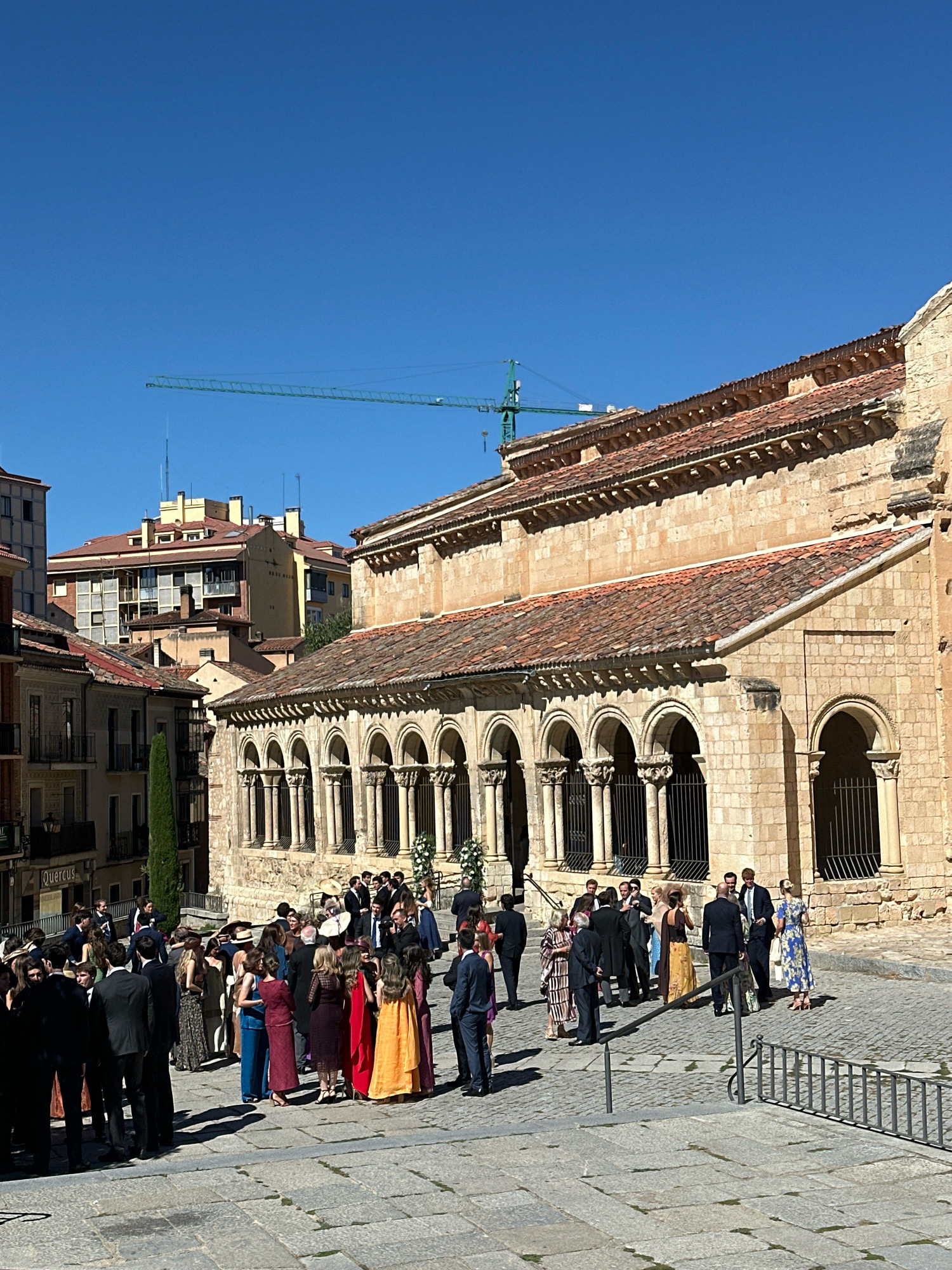 An outdoor daytime medium shot captures a gathering of people dressed formally, appearing to be attending an event such as a wedding, in a cobblestone courtyard. To the right, a stone building with a red tiled roof features an arched portico with multiple columns. A construction crane is visible in the clear blue sky behind apartment buildings in the background. The people are a mix of men in suits and women in dresses. They are standing and conversing in small groups. The scene suggests a European city setting.