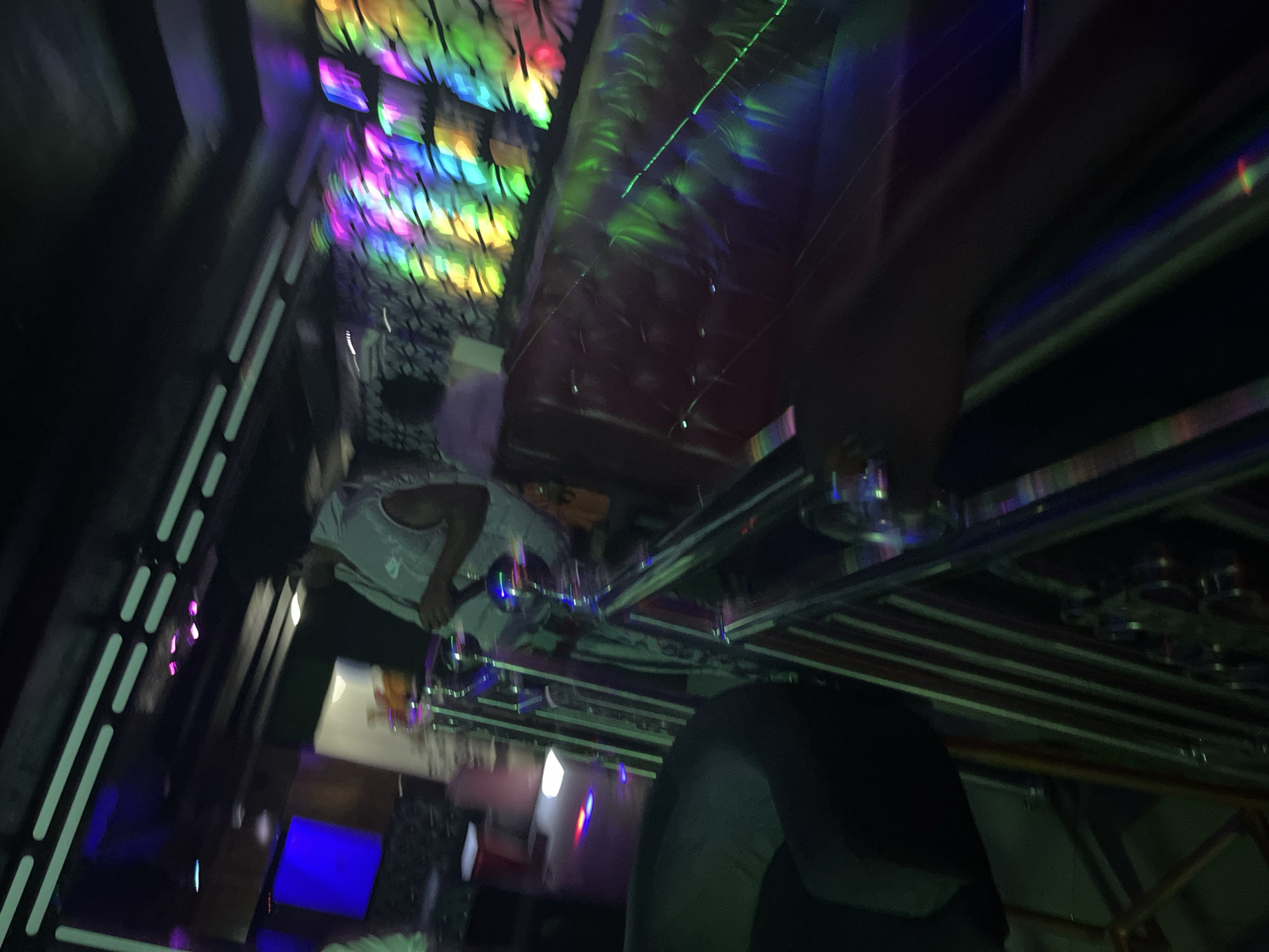 The image depicts an indoor scene, possibly a nightclub or bar, with vibrant and dynamic lighting. In the foreground, there is a red cushioned booth and a metallic railing. In the background, a person wearing a light-colored top with a Nike logo can be seen holding a microphone, suggesting a performance or hosting event. There are also illuminated panels displaying a spectrum of colors, including pink, purple, blue, green, and yellow. The overall ambiance is energetic and nightlife-oriented. The location is identified as Unknown, Unknown.