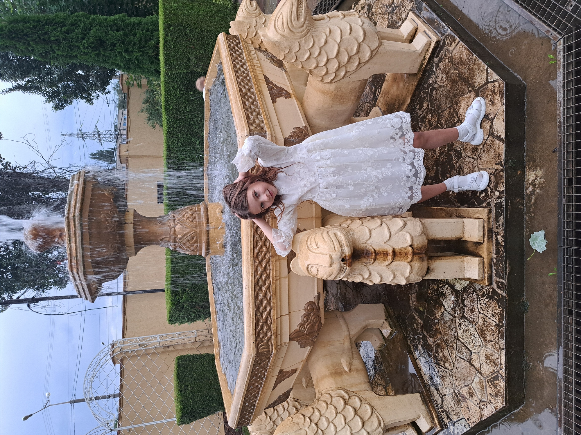A young girl in a white lace dress and white shoes stands in front of a decorative fountain. The fountain has a hexagonal basin supported by four lion-like statues, with water cascading from a central spout. The girl is posing with her hands on her head, looking at the camera. The ground around the fountain is wet and made of stone pavers. In the background, there are trimmed hedges, a wall, an arched trellis, and trees. The sky is overcast.