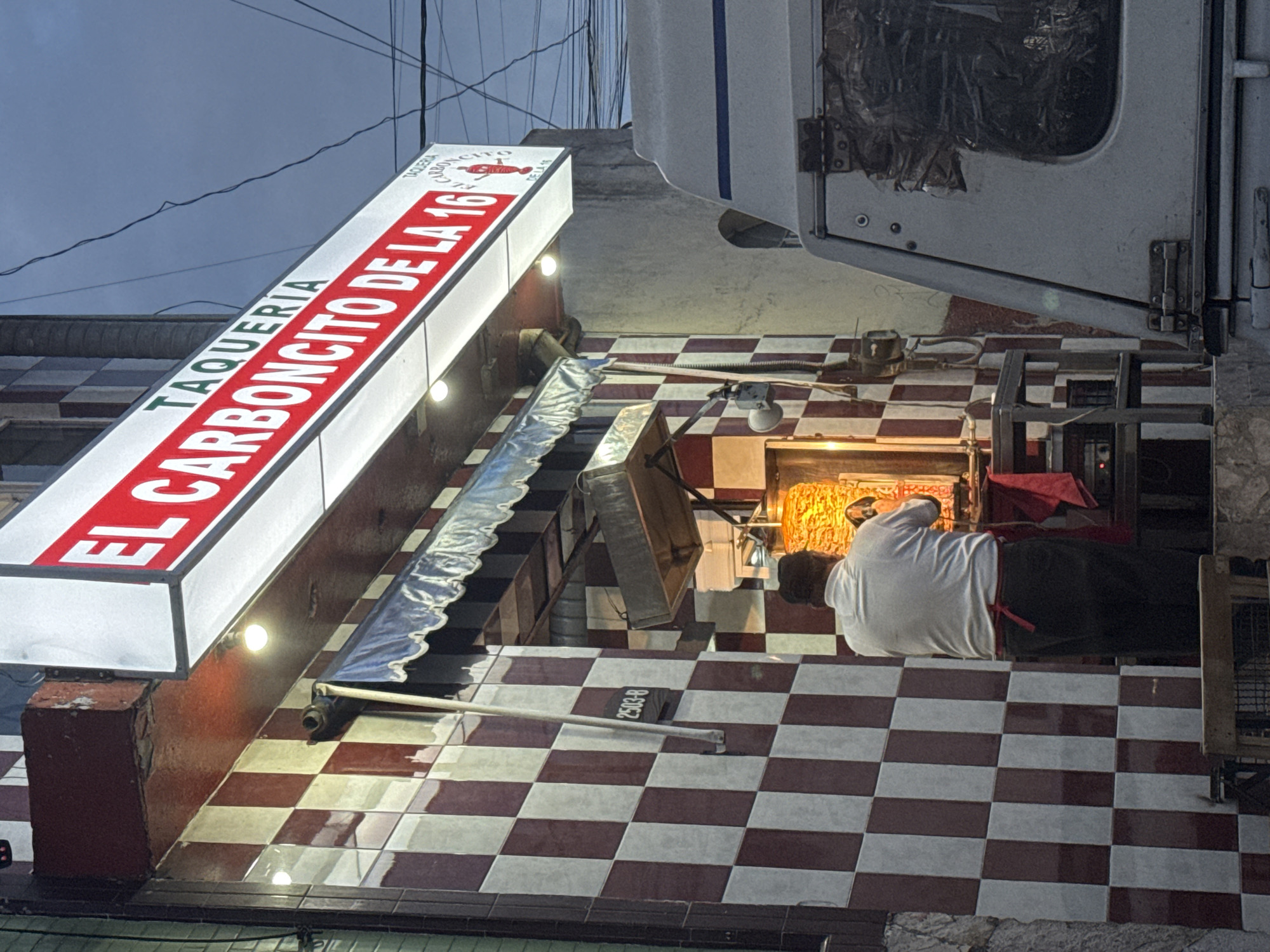 A street-level view depicts a "Taqueria El Carboncito de la 16" storefront in Puebla, Mexico. The red and white sign extends over a tiled awning above a checkerboard red and white tile floor. A cook in a white shirt and dark pants, with a red apron is working at a meat grill. Some cooking tools are on the checkerboard red and white tile floor near the grill. A white panel truck with an open back is parked at the curb. Overhead electrical wires are visible.