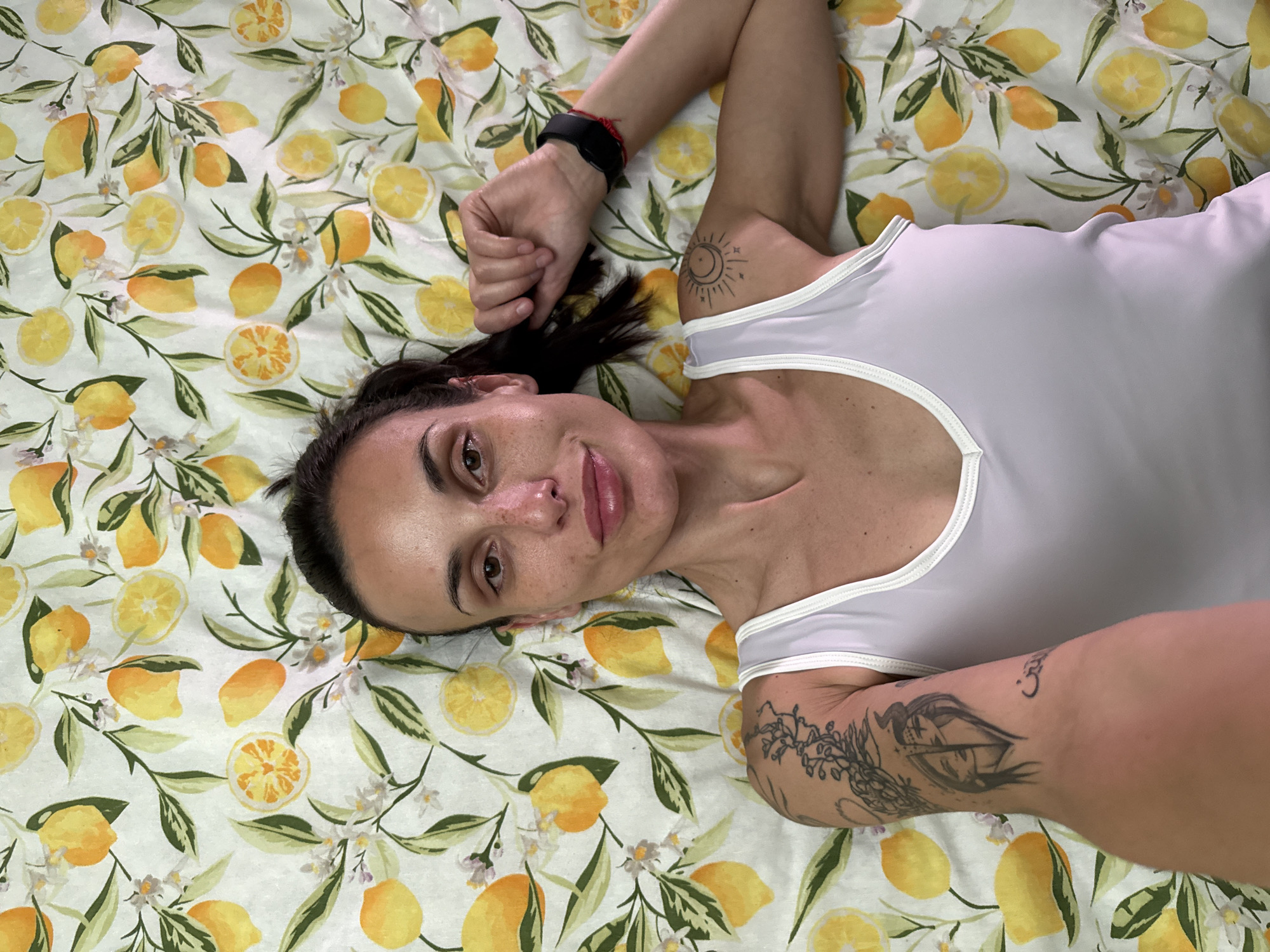 A woman lies on her back on a lemon-patterned fabric. She is wearing a white tank top. Her arms are raised above her head, and tattoos are visible on both arms. She wears a black watch or fitness tracker on her left wrist. Her dark hair is pulled back from her face. The background is entirely filled with the lemon-print fabric.
The provided information does not include the location of the image in Pleven, Bulgaria.