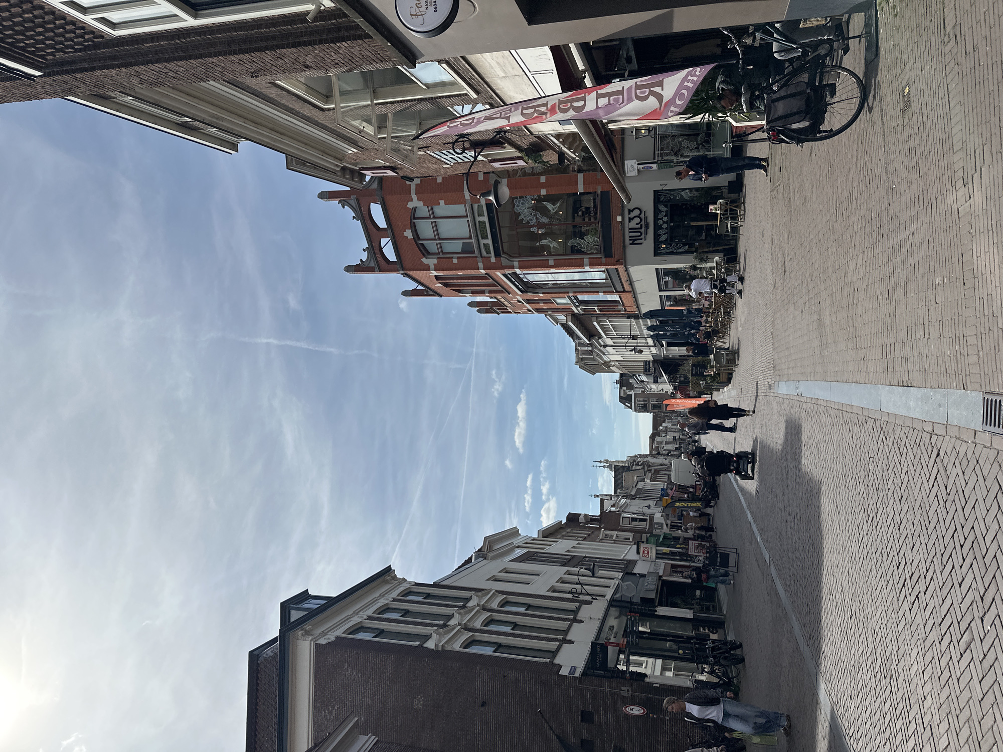 The image shows a street in Amersfoort, Netherlands. It is a narrow pedestrian zone paved with brick. Buildings flank both sides of the street. Several shops and cafes are visible, with awnings and signage. People are walking along the street and sitting at outdoor tables. A bicycle is parked on the right side of the street. The sky is blue with some clouds.