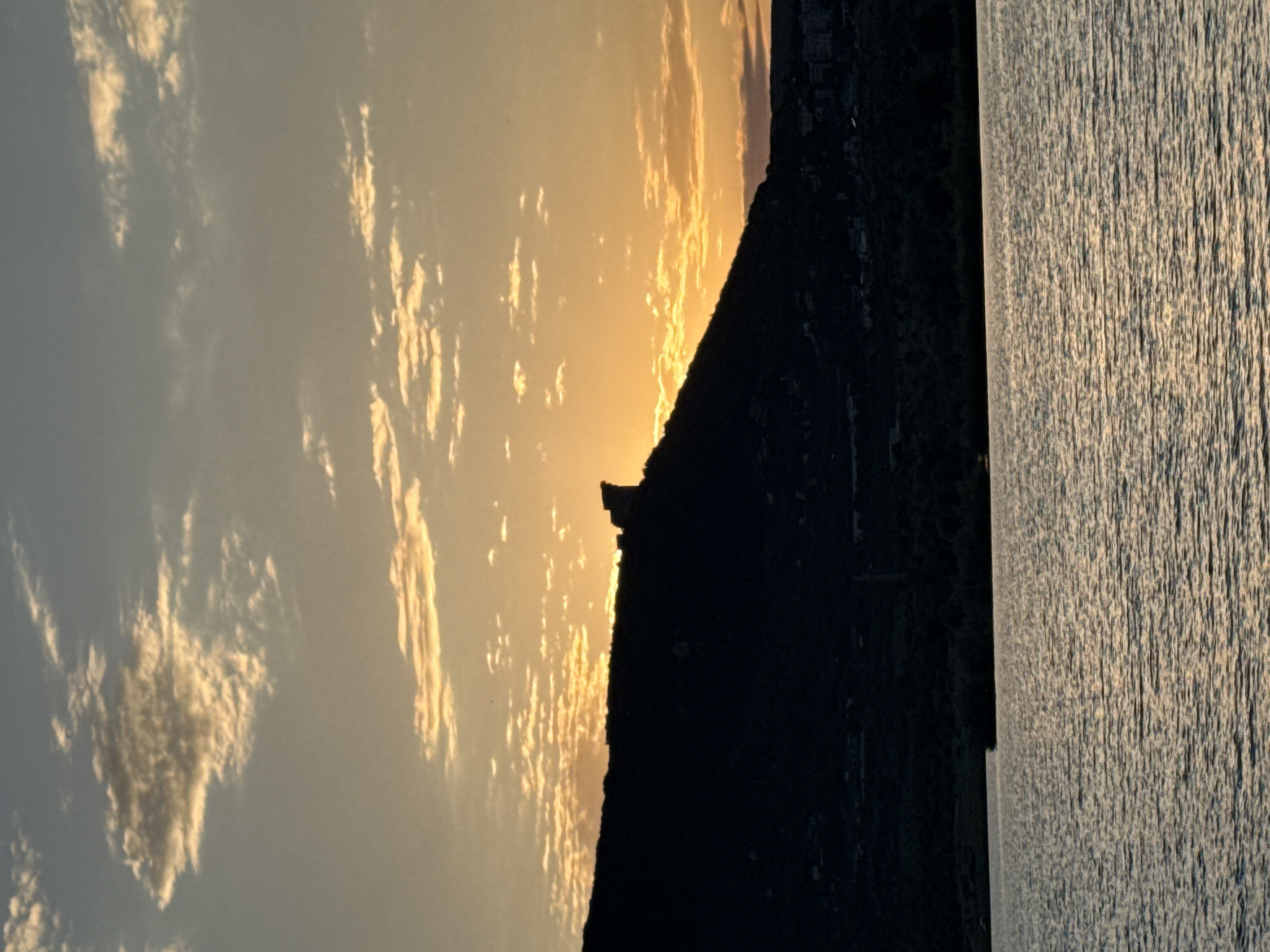 The image depicts a silhouette of a rocky prominence on a hillside, potentially the Shumen Fortress, against a sunset sky. The sky is filled with scattered clouds illuminated by the setting sun, casting an orange and yellow glow. The foreground features a body of water, likely a lake or river, reflecting the colors of the sky. The hillside is covered in dark vegetation, and hints of buildings are visible on the lower slopes. The overall scene is tranquil and scenic, capturing the natural beauty of the area surrounding Shumen, Bulgaria.