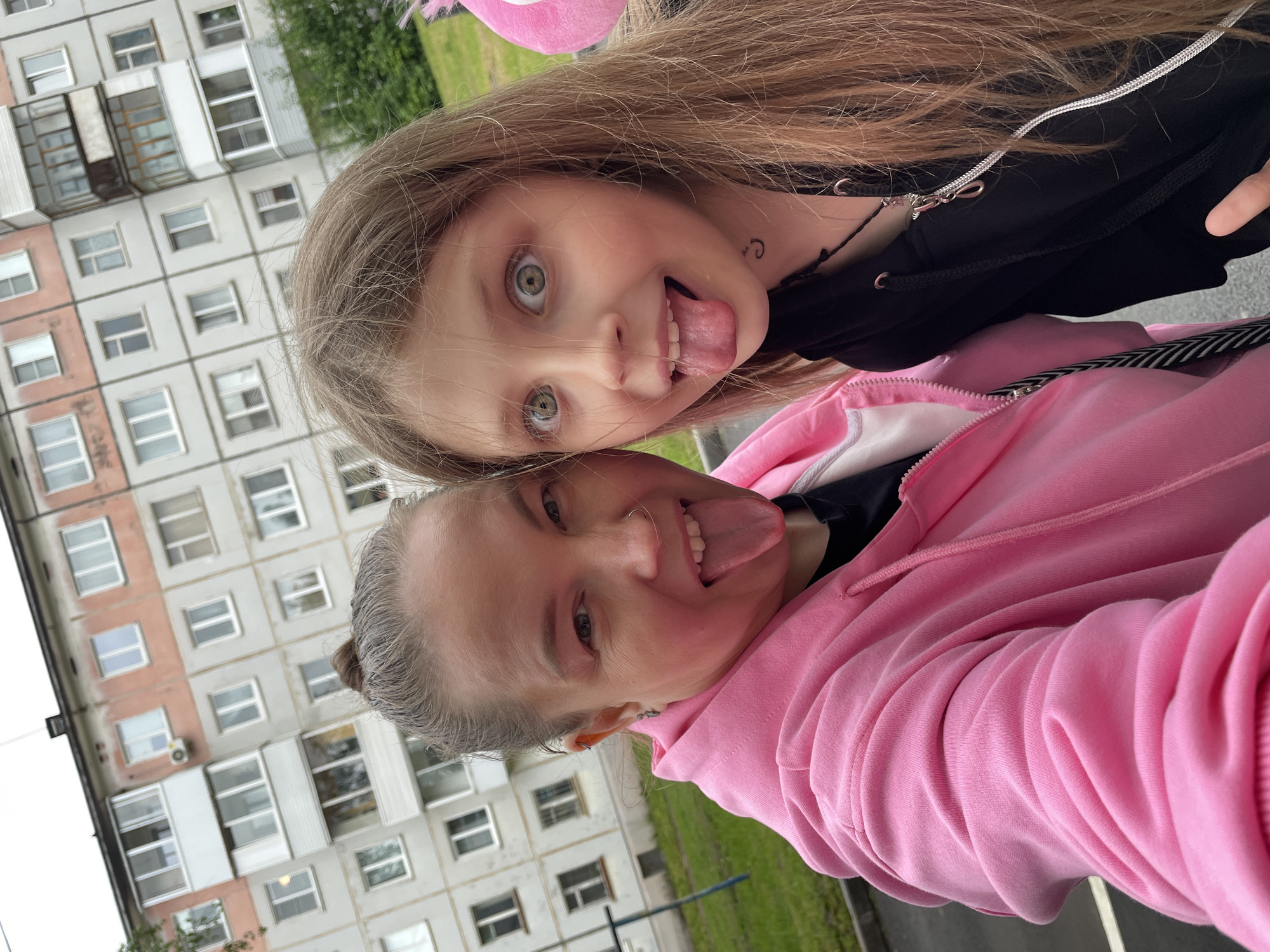 Two individuals, a younger female and an older female, are posing closely together for a photograph, both sticking out their tongues. The younger female is positioned above the older female. The younger female has long blonde hair and is wearing a black top. The older female is wearing a pink hooded sweatshirt. The background reveals an apartment building in Izhevsk, Russia.