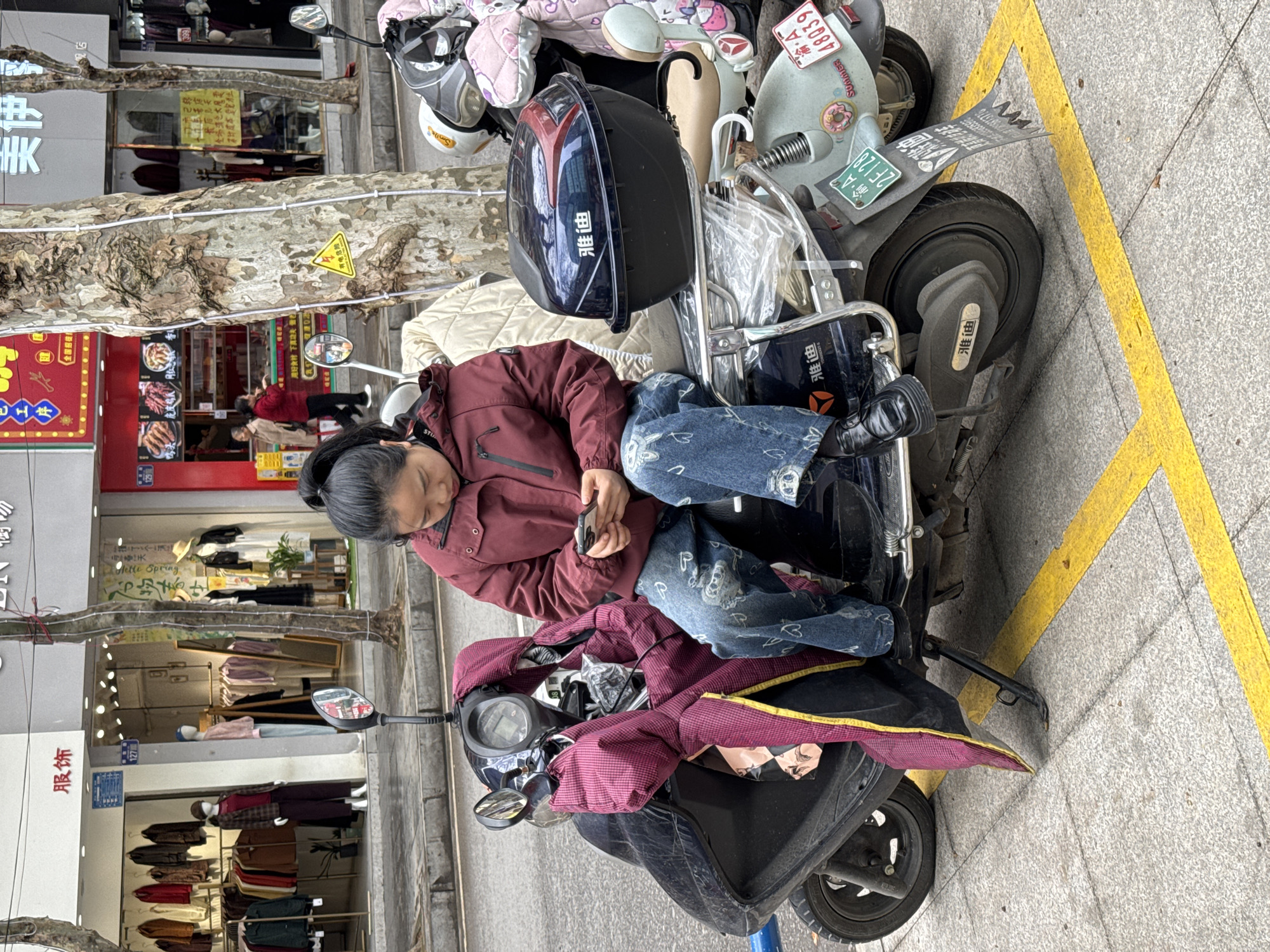 The image shows an elderly woman sitting on a black electric scooter parked on a sidewalk in Qianjiang, China. She is wearing a maroon jacket and jeans, and looking at a mobile phone in her hands. The scooter is parked next to a yellow no-parking zone on the sidewalk. Several other scooters and mopeds are parked behind it. The street is lined with shops and restaurants. The street address "127" is visible. A sign reads "Helle Spring."
