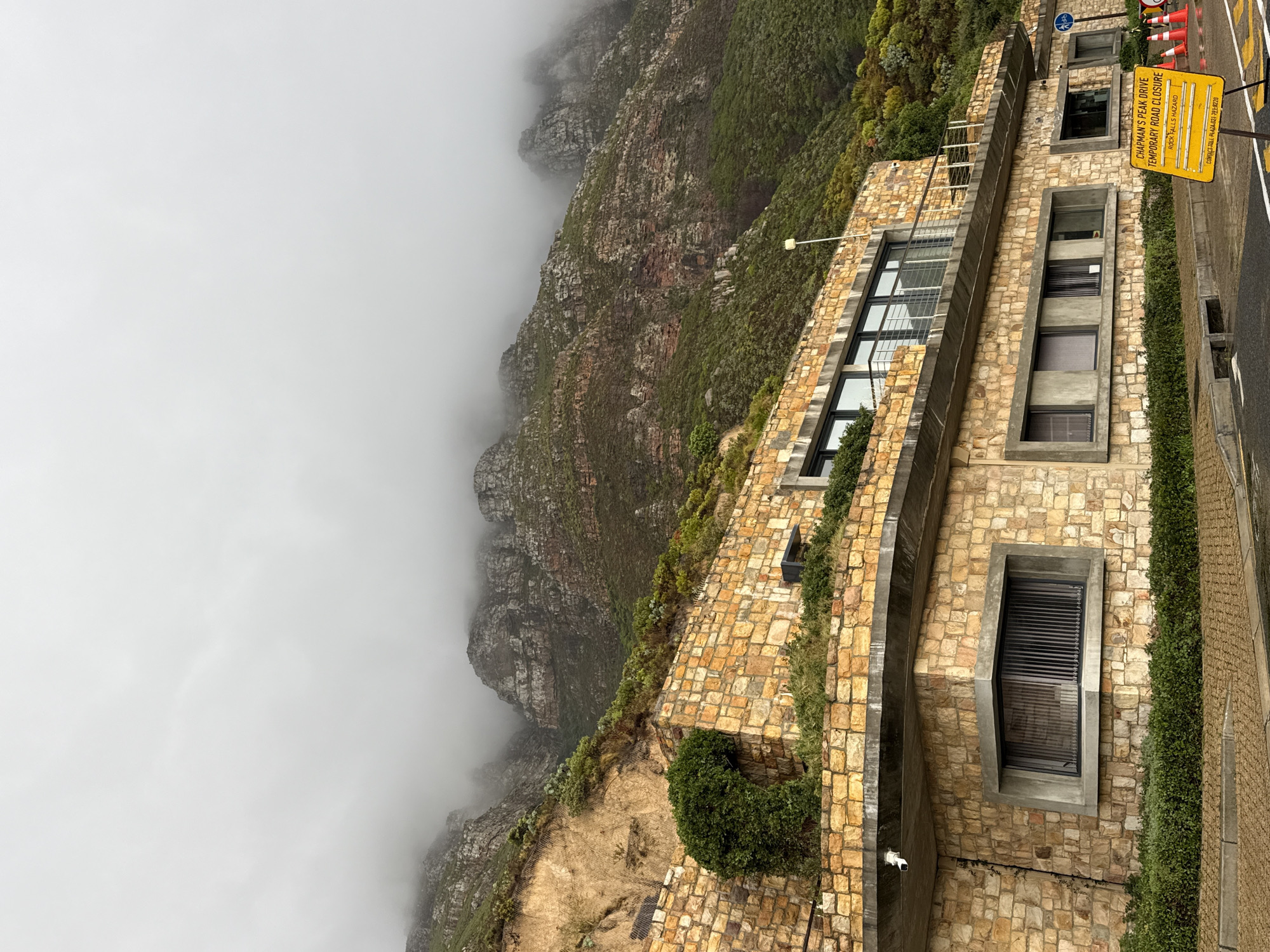 The image showcases a section of Chapman's Peak Drive, a scenic coastal road near Hout Bay, South Africa. The road is temporarily closed due to a rock falls hazard, indicated by a yellow warning sign and orange traffic cones. A stone wall structure with modern windows runs along the road, blending with the adjacent rocky cliffs shrouded in mist. The cliffside vegetation is visible along the edge. The overall scene depicts a combination of natural landscape and man-made architecture, emphasizing the potential dangers of the environment.