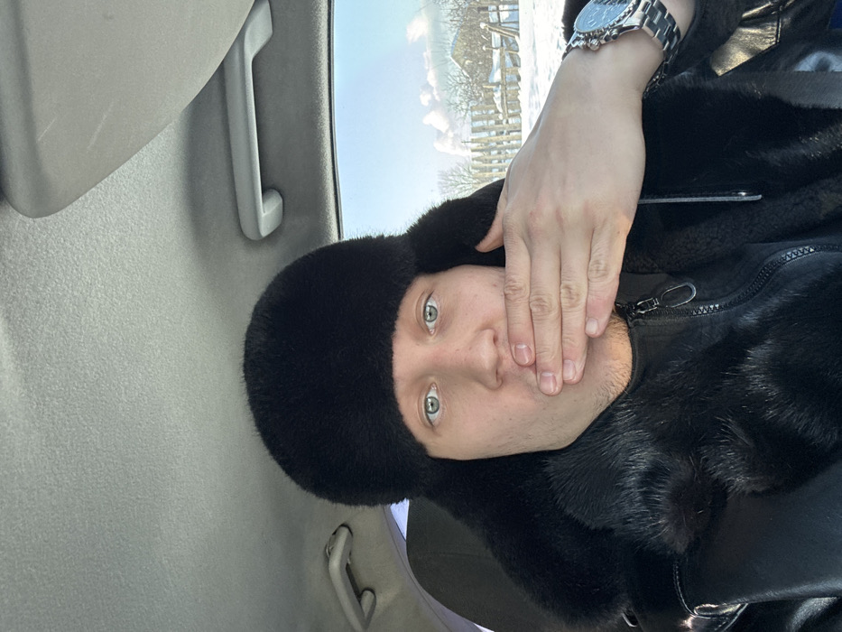 A male individual, wearing a black fur ushanka hat and a black, fur-trimmed jacket, is seated inside a vehicle. His light-colored eyes are visible, and his right hand, adorned with a silver-toned metallic wristwatch, covers his mouth. The vehicle's grey headliner, a grab handle, and a sun visor are visible in the interior. Through the vehicle's window, a snowy winter landscape in the city of Ольховка, Russia, is visible. The exterior scene features snow-covered ground, bare trees, a wooden fence, and a distant building under a clear blue sky with sparse clouds.