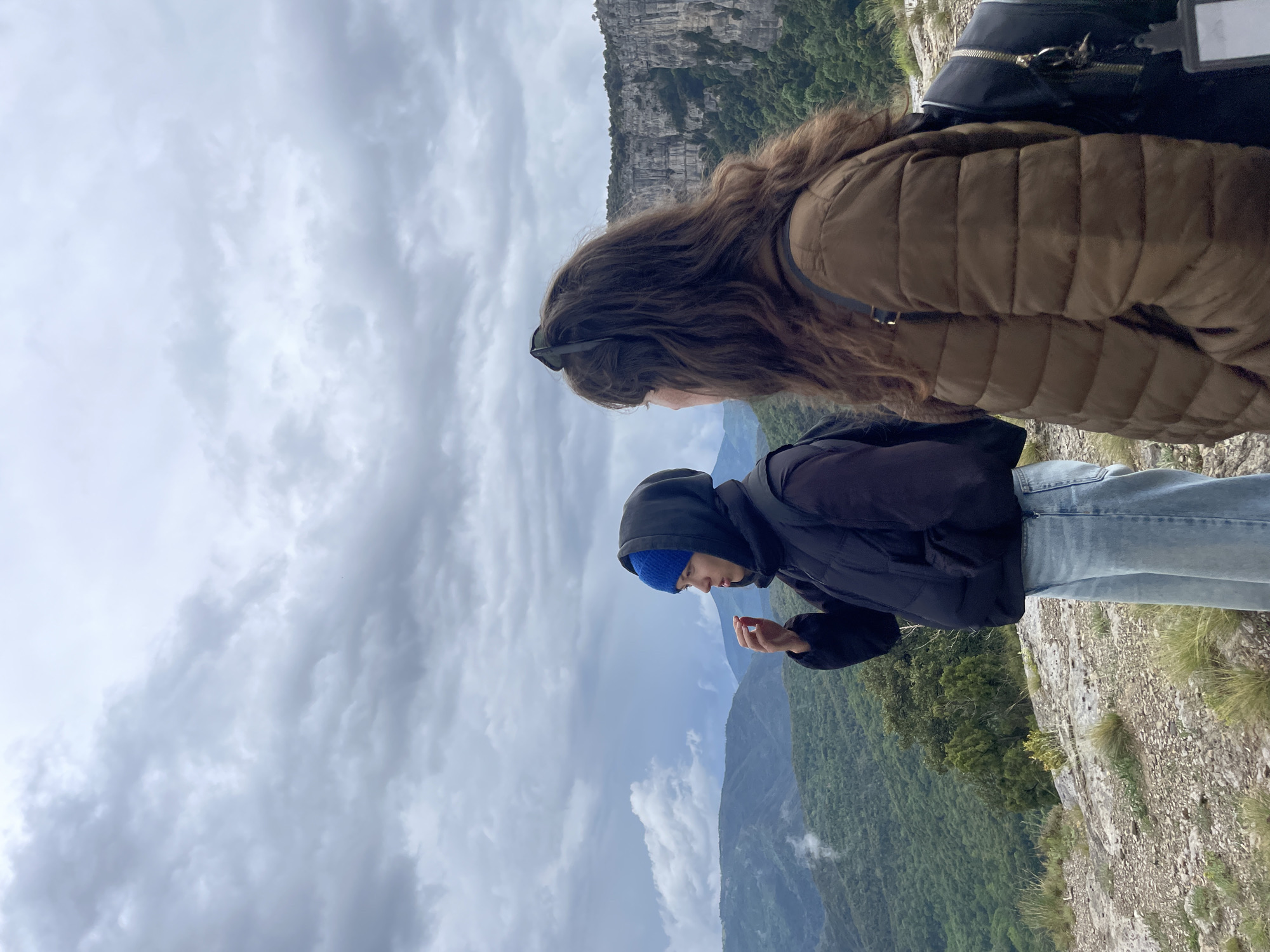 Two individuals stand on a cliff's edge overlooking a valley in Rupit i Pruit, Spain. The foreground shows a person with long, wavy brown hair wearing sunglasses, a brown quilted jacket, and a black backpack. In front of them is another person wearing a black hooded jacket over a blue knit hat, light blue jeans, and holding an unidentified object in their hand. The valley contains green vegetation and stretches into the distance, where mountains and clouds are visible. The sky is overcast.