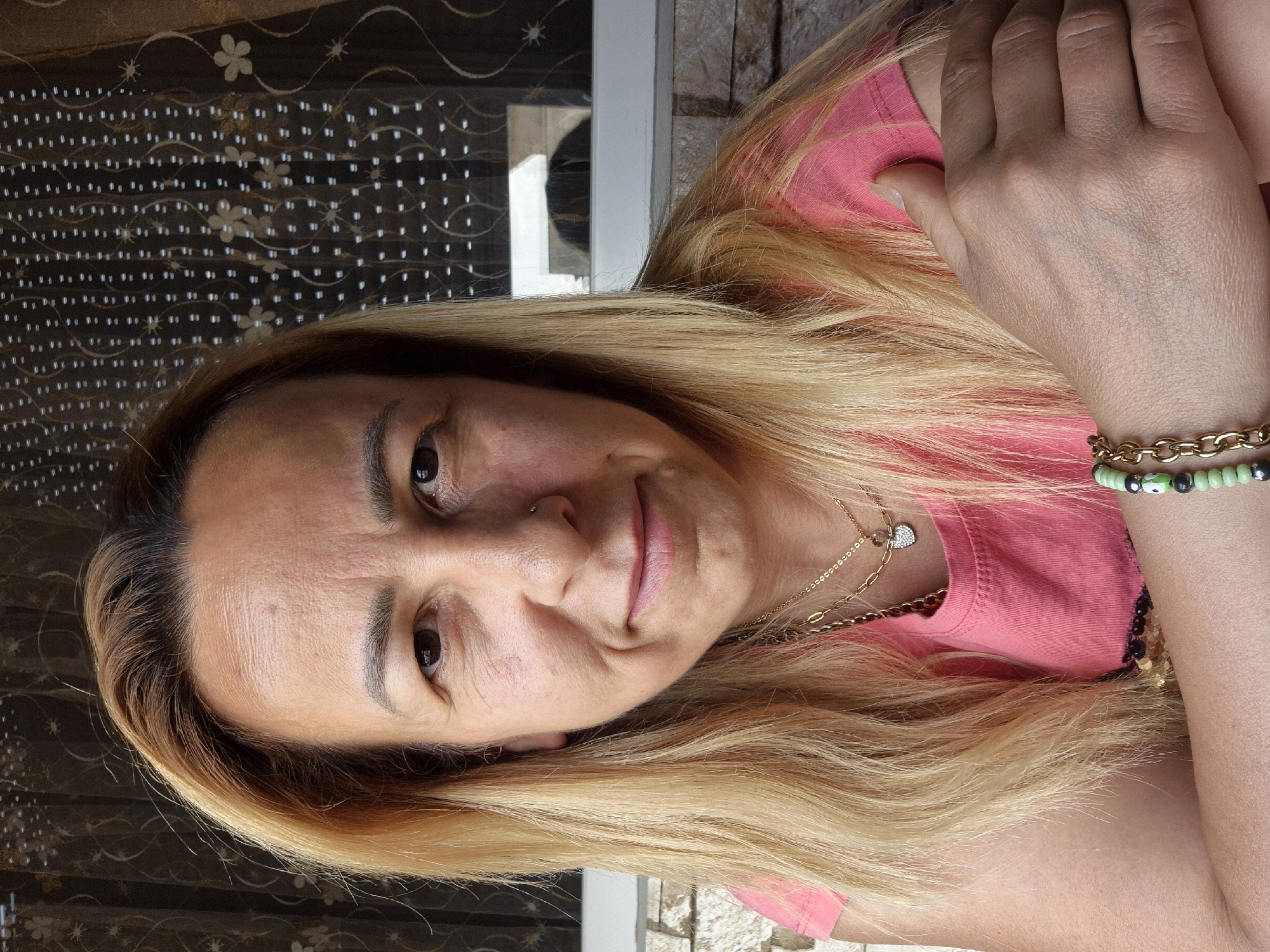 A close-up, head-and-shoulders selfie features a woman with medium-length blonde hair, a pink t-shirt, and two necklaces. She has a nose ring and is smiling slightly. Her arms are crossed in front of her chest, and she is wearing a green beaded bracelet on her left wrist. The background is a window with a patterned curtain. The setting appears to be outdoors, possibly on a patio or balcony, with a stone wall visible in the lower portion of the image.