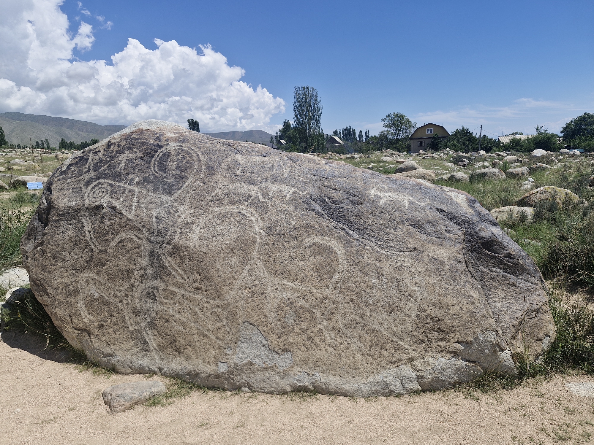 The image shows a large, rough-textured boulder with petroglyphs carved into its surface. The boulder is situated in a dry, grassy landscape with scattered smaller rocks. In the background, there are some trees, houses, and distant mountains under a bright blue sky with scattered clouds. The petroglyphs on the boulder appear to depict abstract shapes and possibly animal figures. The scene is illuminated by daylight. This location is likely part of the Cholpon-Ata petroglyph site in Kyrgyzstan.