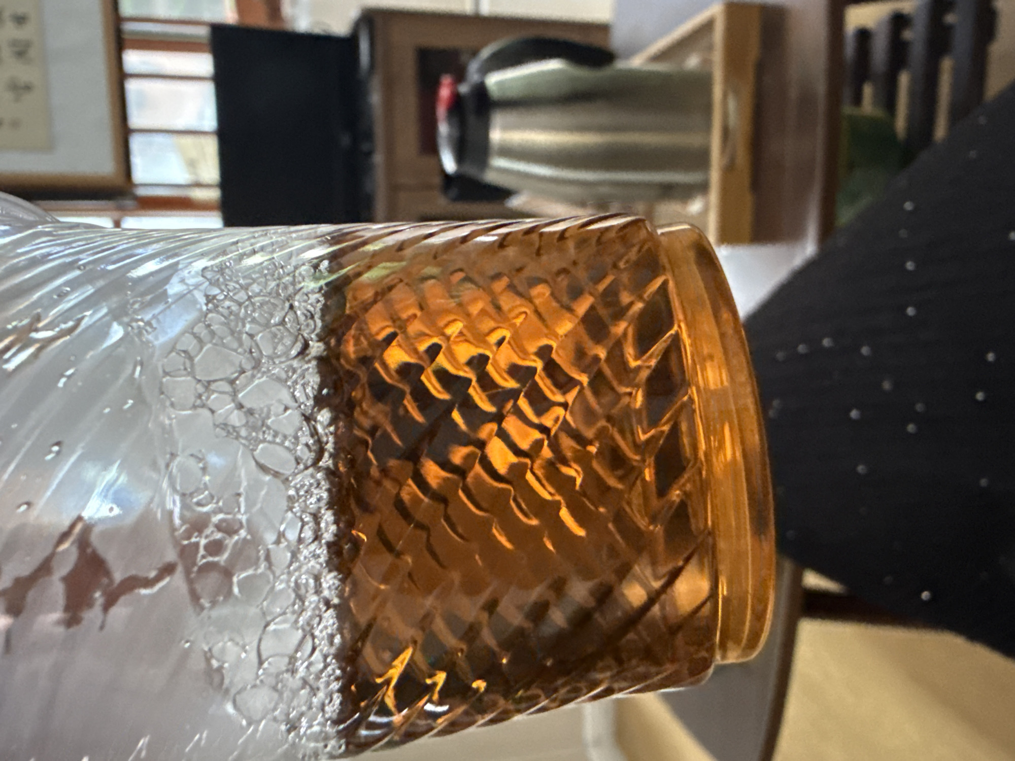 An amber liquid fills a clear, textured plastic cup, with a layer of bubbles at the top. The cup is held at an angle, showing the textured pattern of the plastic. In the background, a stainless steel thermal carafe with a black lid and red accent sits on a wooden tray on a wooden table. Further in the background, a dark television screen is visible behind a wooden cabinet, with windows and a framed artwork on the wall above. The scene appears to be indoors, possibly in a home or common area. There are no discernible people or specific locations within Huizhou, China that can be identified from this image.