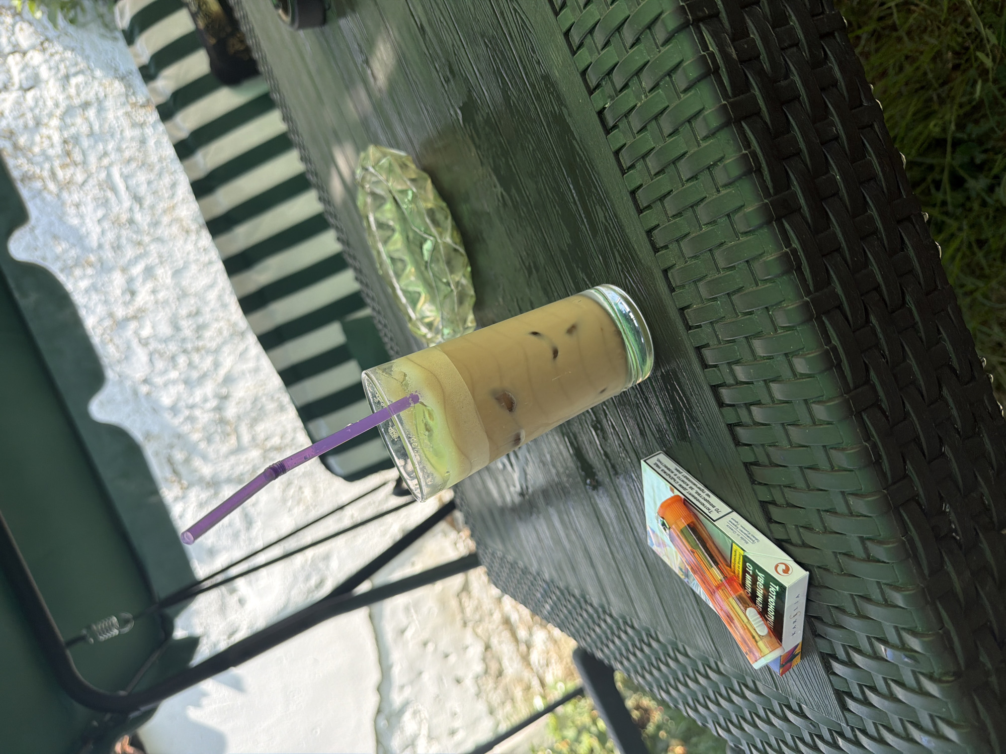 The image depicts an outdoor scene, likely in Razhevo Konare, Bulgaria, on a table. A glass of iced coffee with a purple straw sits prominently on the table. To the left of the coffee is a pack of cigarettes, with an orange lighter resting on top of it. An ashtray is visible to the right of the coffee. The table appears to be made of dark, woven material. In the background, there is a swing with a green canopy and striped cushions, suggesting a relaxed outdoor setting. The overall lighting indicates it is daytime. There are no people visible in the image.