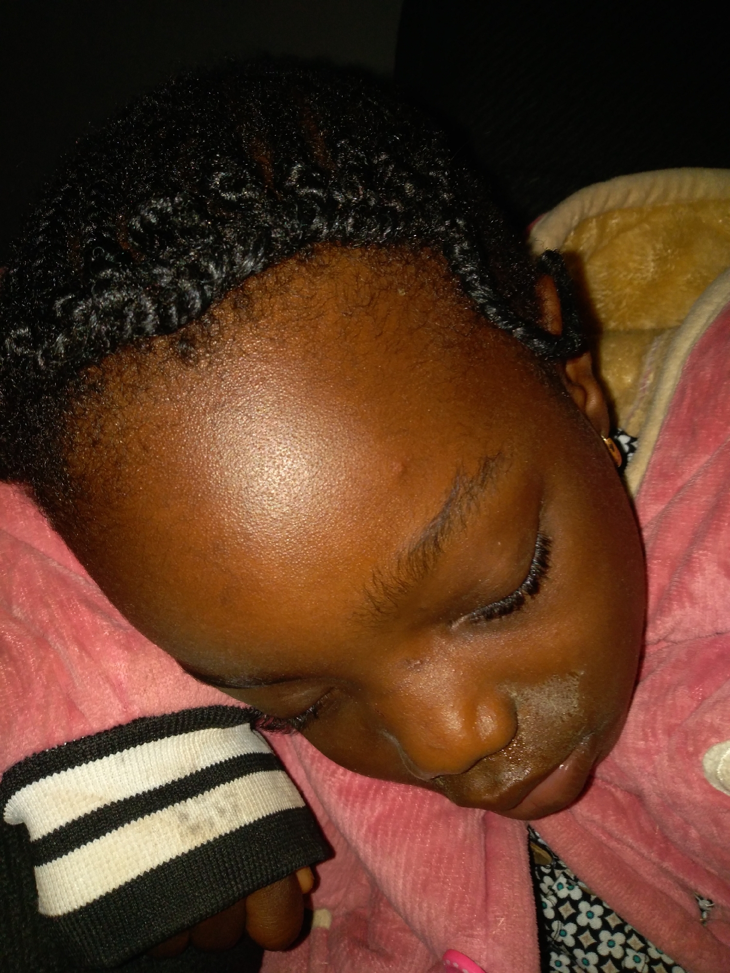 A close-up, overhead shot captures a young Black child sleeping. The child's head is turned to the left, with their face resting on a pink, plush fabric. Their eyes are closed, and their eyelashes are visible. The child's hair is black and styled in small, textured braids. They are wearing a pink hooded jacket with a white and black striped cuff on the sleeve. Part of a yellow blanket is visible behind the child's head. The background is dark and out of focus. The scene appears to be indoors, with no specific city or environmental details discernible.