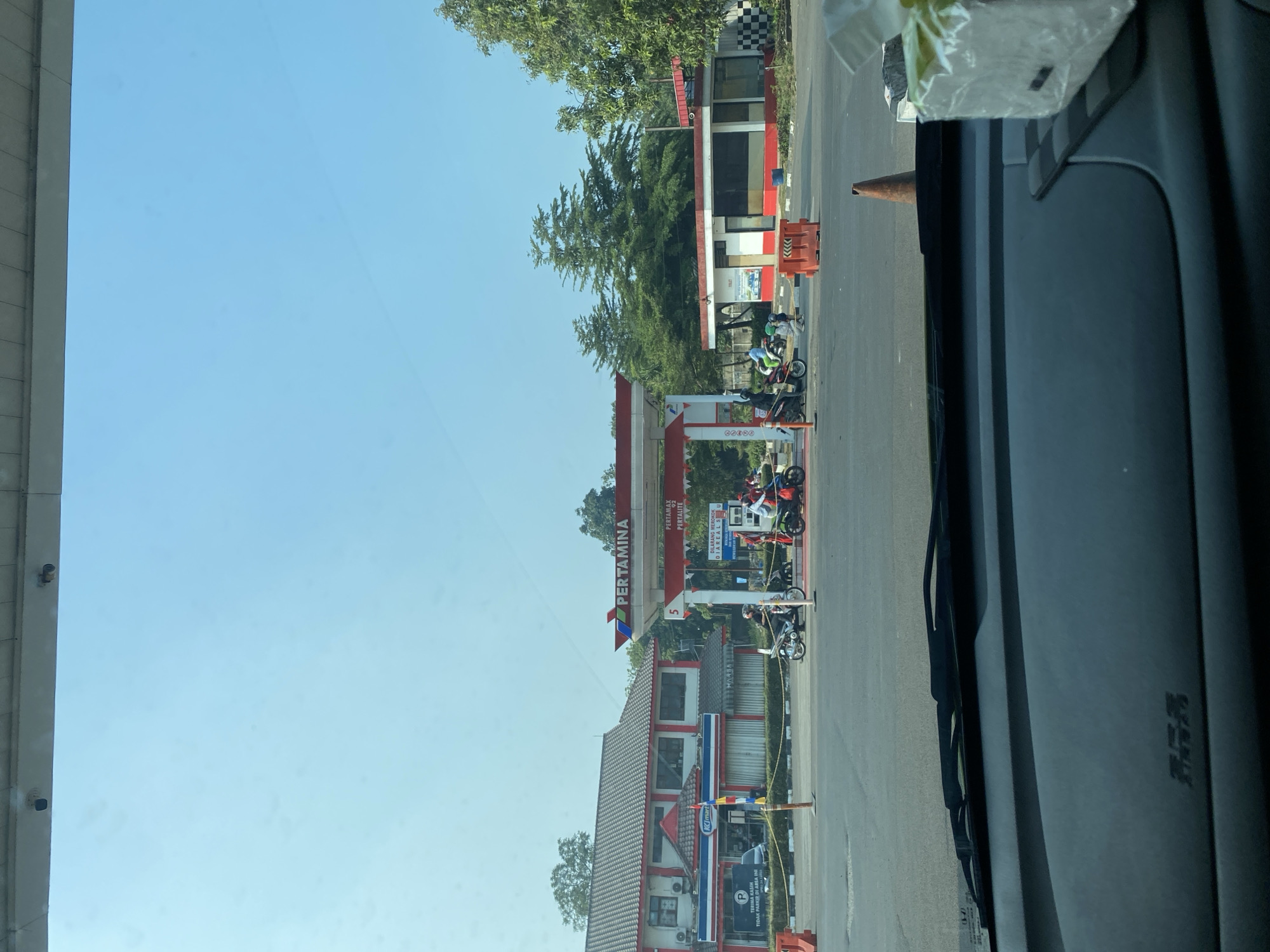 The image captures a view from inside a vehicle at a Pertamina gas station in Gunung Putri, Indonesia. The gas station features a prominent red and white canopy with the Pertamina logo. Several motorcycles are present, with some drivers appearing to be refueling. A K-Mart store is visible next to the gas station. The sky is clear and blue, suggesting a sunny day. The foreground shows the car's dashboard and windshield wipers. The scene depicts a typical day at a bustling gas station in Indonesia.