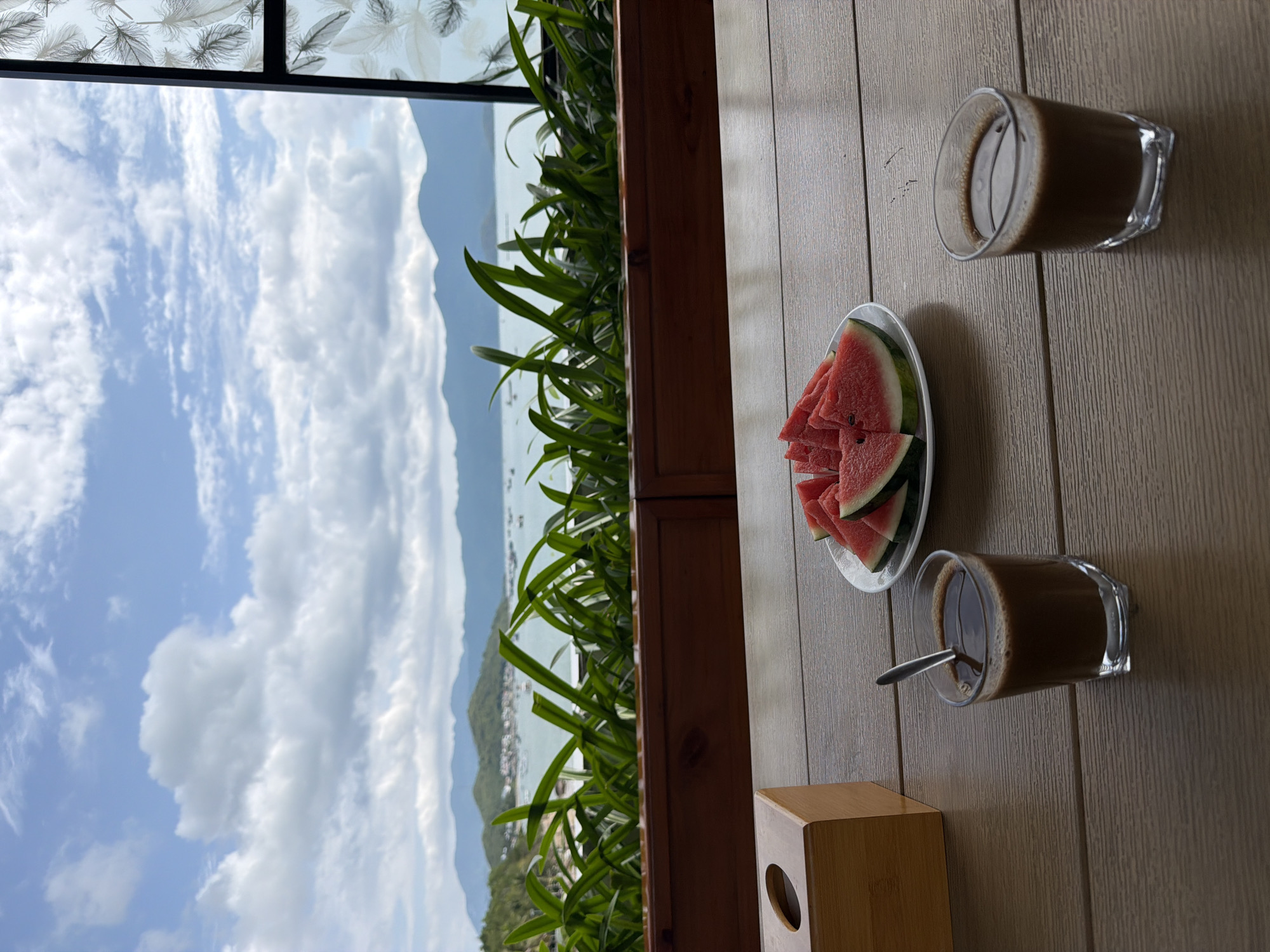 A view of the coastal Bac Nha Trang Ward, Vietnam, is observed from a high vantage point. The image features a window perspective. Outside, a cloudy sky and a seascape are visible, with a suggestion of hilly terrain. Indoor, there is a wooden table with a plate of sliced watermelon, two glasses of coffee, and a tissue box. A green plant is also present near the window.