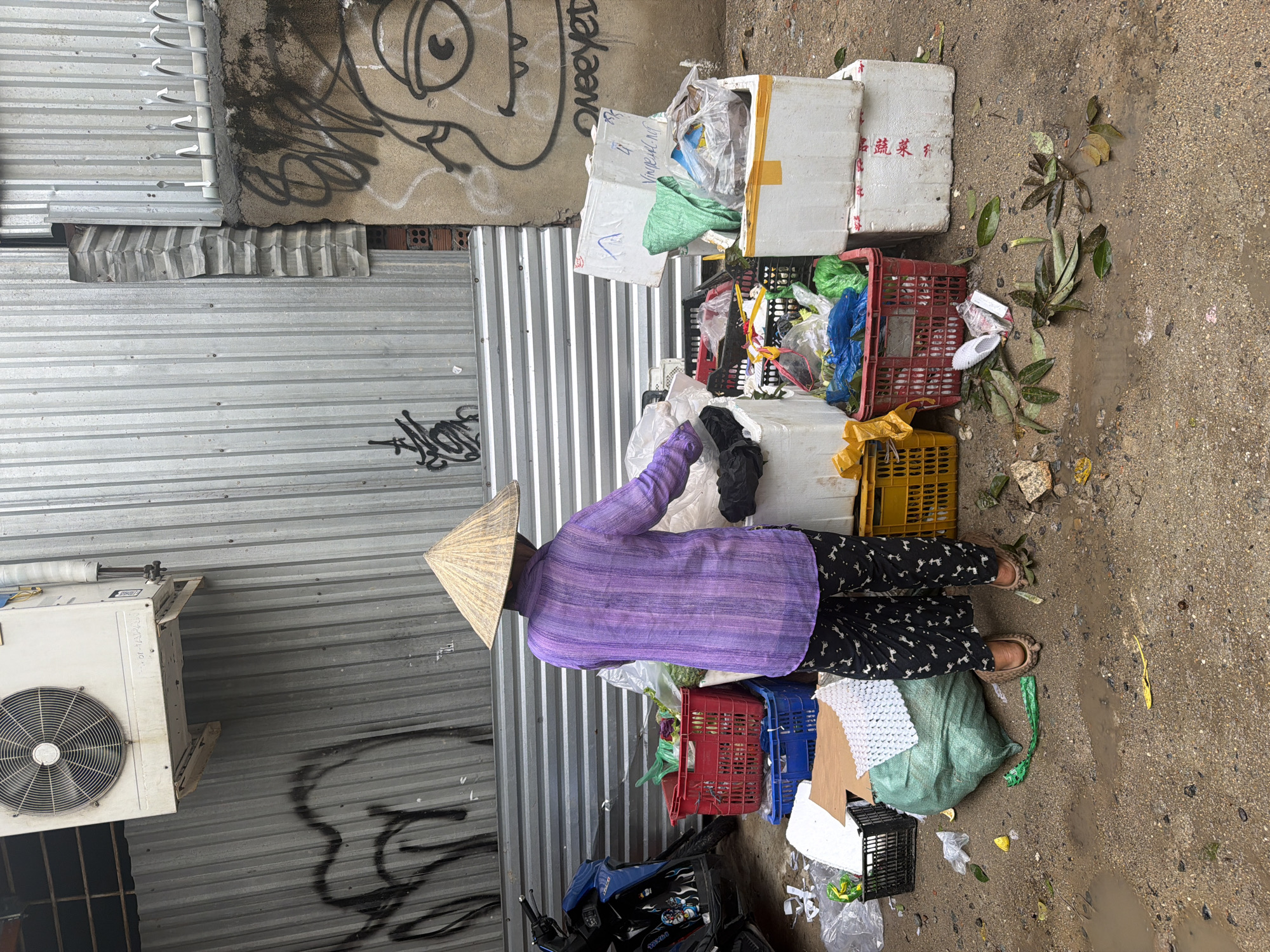 The image depicts a person wearing a traditional conical hat and a purple shirt, standing amidst a collection of crates and styrofoam boxes overflowing with various items, likely recyclables or salvaged goods. The setting is an alleyway in what appears to be an urban environment, evident from the corrugated metal walls covered in graffiti and an air conditioning unit mounted on one wall. A motorcycle is parked nearby, partially obscured by a blue tarp. The ground is wet and littered with debris, suggesting recent rain or damp conditions. The location is identified as Bac Nha Trang Ward, Vietnam. The individual appears to be sorting through the items, possibly collecting materials for recycling or reuse. The overall scene suggests informal waste management or scavenging activities within the urban landscape.