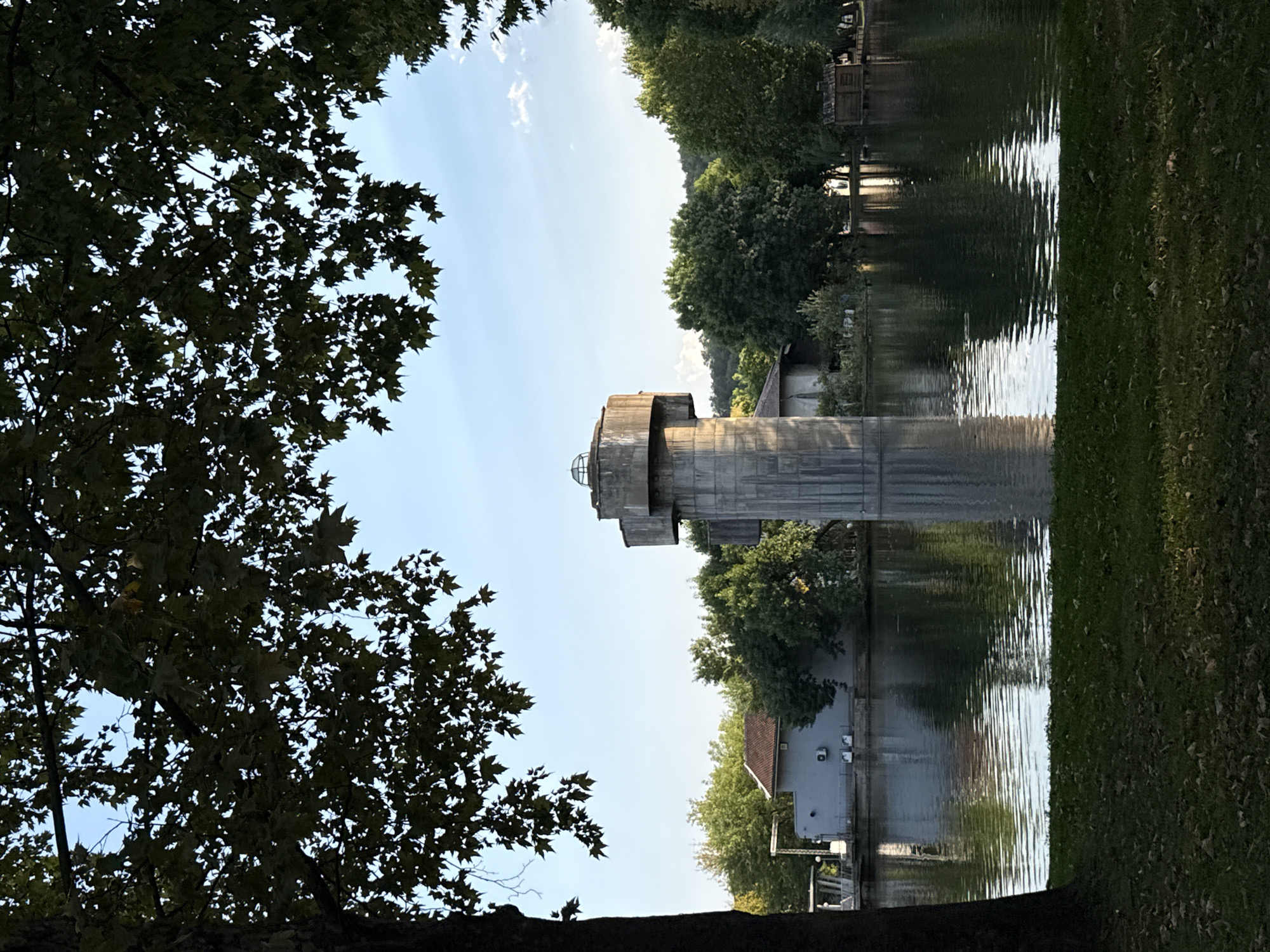 The image depicts a tranquil scene at the lake in Montana, Bulgaria. In the foreground, a lush green lawn with fallen leaves is visible, leading to the calm waters of the lake. Dominating the center of the frame is a striking water tower, a concrete structure with a distinctive cross-shaped top, standing tall in the middle of the lake. Its reflection shimmers on the water's surface.
Across the lake, various buildings and dense greenery are scattered along the shoreline. On the left, a light-colored building with a tiled roof is partially obscured by trees. A small wooden structure, resembling a shed or hut, sits on stilts on the right side of the lake, also surrounded by trees. The water is calm, with gentle ripples reflecting the clear blue sky, which is dotted with a few wispy clouds. Overhanging branches of a large tree in the foreground frame the top of the image, adding depth and a sense of natural enclosure to the scene. The overall atmosphere is peaceful and serene, suggesting a beautiful day in a park-like setting.
