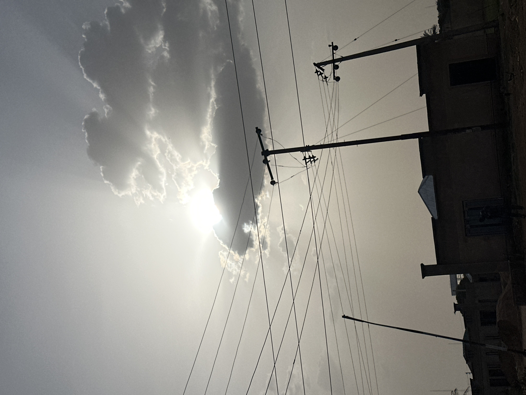 This monochrome image captures a scene with power lines cutting across the sky, partially obscured by clouds. The sun is positioned behind the clouds, creating a bright halo effect. To the right, buildings are visible, some with visible windows and architectural details. The scene is presumed to be in Bukuru, Nigeria. There are no visible people or actions depicted. The environment suggests a town with electrical infrastructure.