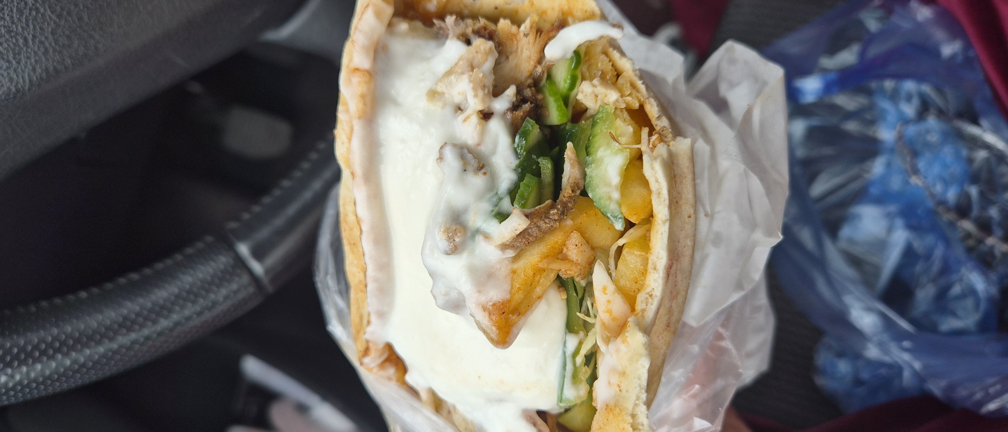 A shawarma is presented in a plastic wrapper. Inside the pita bread, there is white sauce, green peppers, yellow fries, and meat. To the left of the shawarma is the steering wheel of a car. To the right of the shawarma is a blue plastic bag and a piece of red cloth. It cannot be determined from the image whether this image was taken in Plovdiv, Bulgaria. There is no information about people, actions, or interactions.