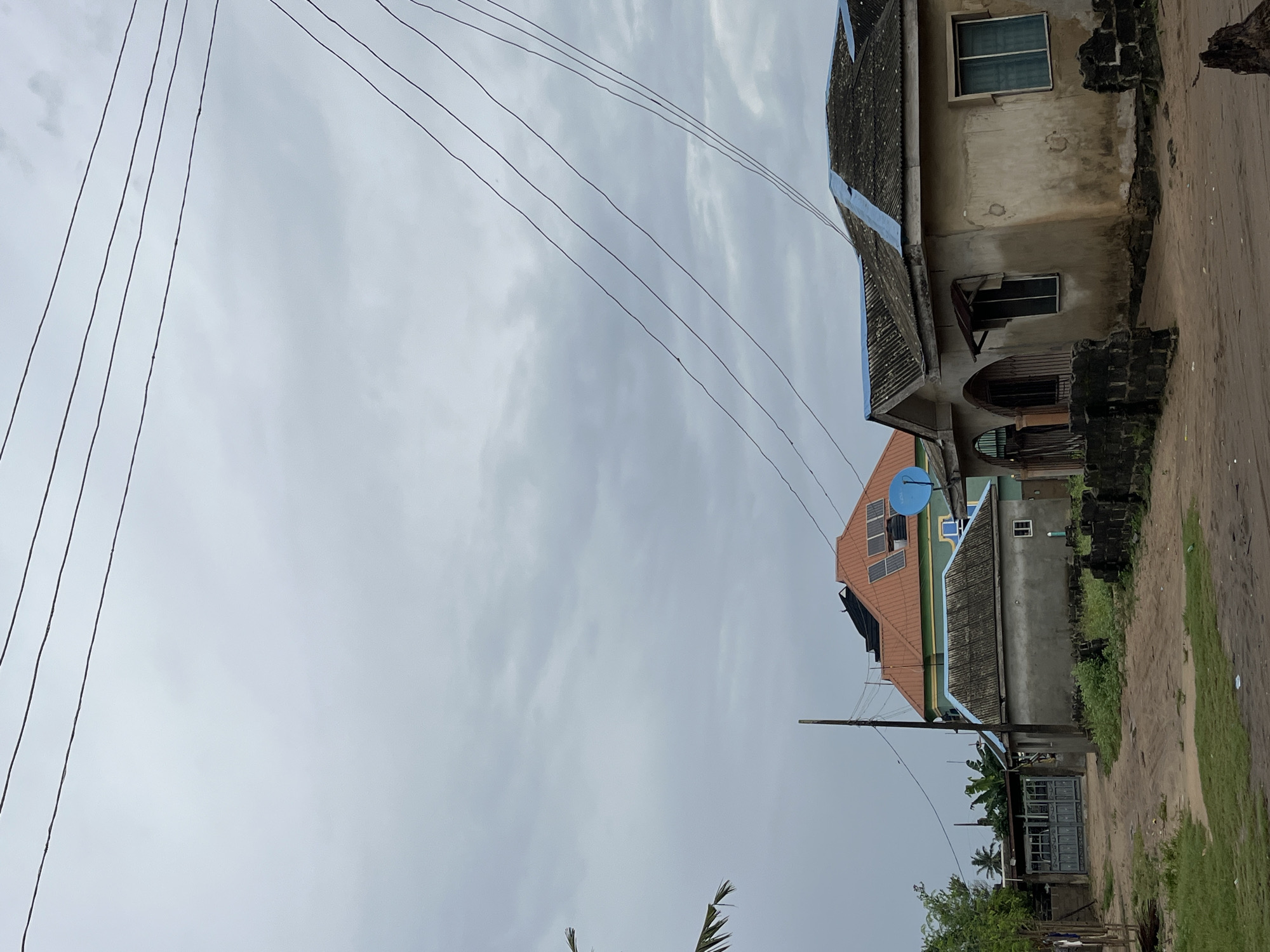 The image shows a rural scene in Badagry, Nigeria. In the foreground, a dirt road is visible, with some sparse green grass and a low stone wall. To the right, a light-colored building with a corrugated metal roof and a blue trim stands partially in shadow. The building has a window and an arched entrance with metal gates. Further back, a larger building with a red-tiled roof and solar panels is visible. To the left of this building is a grey structure with a corrugated roof and a blue trim, and a large grey metal gate. Trees and palm fronds are scattered throughout the scene. Overhead, multiple power lines crisscross the cloudy sky. The overall impression is one of a modest, lived-in community.