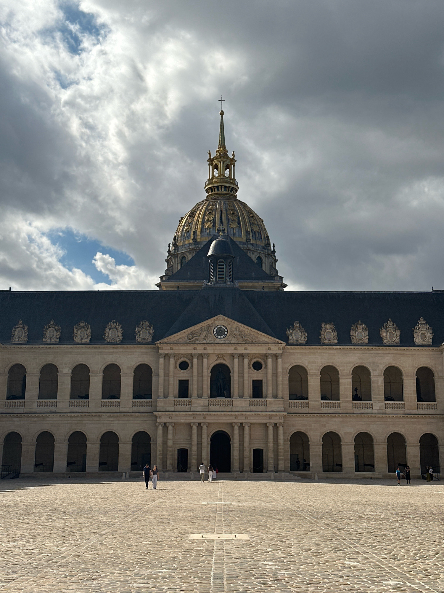 This is a photograph of the exterior of the Hôtel des Invalides in Paris, France. The building features a large, ornate golden dome topped with a cross. The facade is made of light-colored stone and is characterized by rows of arched windows and classical architectural details. In the foreground, a large cobblestone courtyard is visible, with a few people walking across it. The sky is filled with dramatic, cloudy formations.