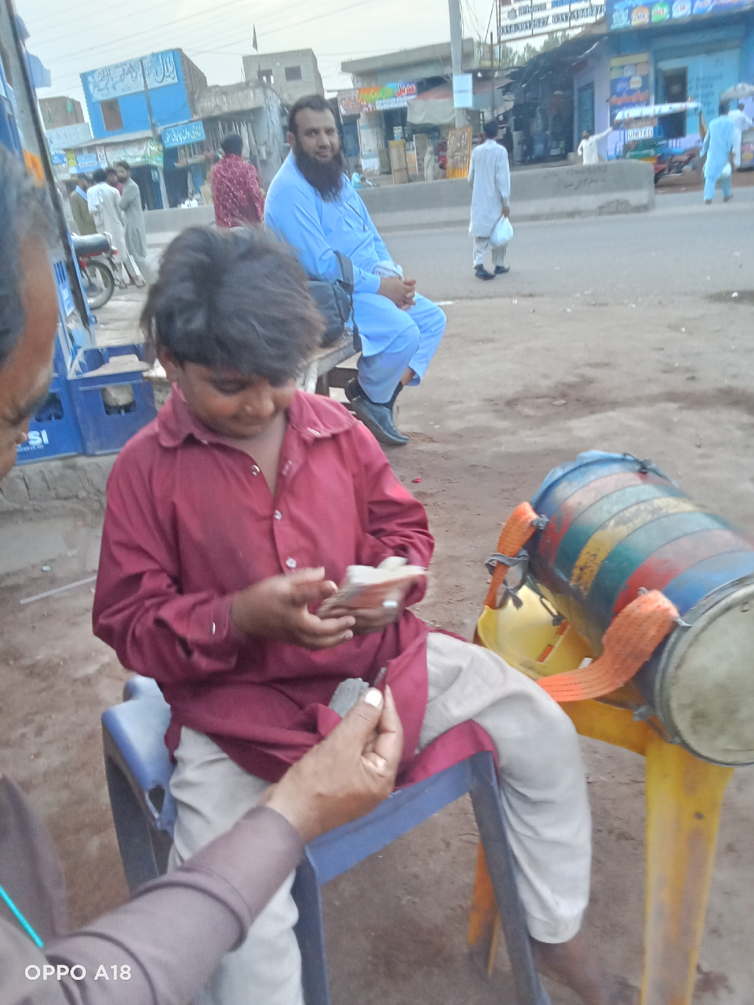 A child in a maroon shirt sits on a blue plastic chair, counting money. A man's hand, seemingly an adult, offers a bill. Next to the child is a colorful drum resting on a yellow plastic chair. Behind the child, a man with a beard is seated, wearing a blue outfit. Further back, there's a street scene with people walking and storefronts visible. The photo was taken in a city, with location details "Unknown, Unknown." A motorcycle parked on the left side can also be seen.
