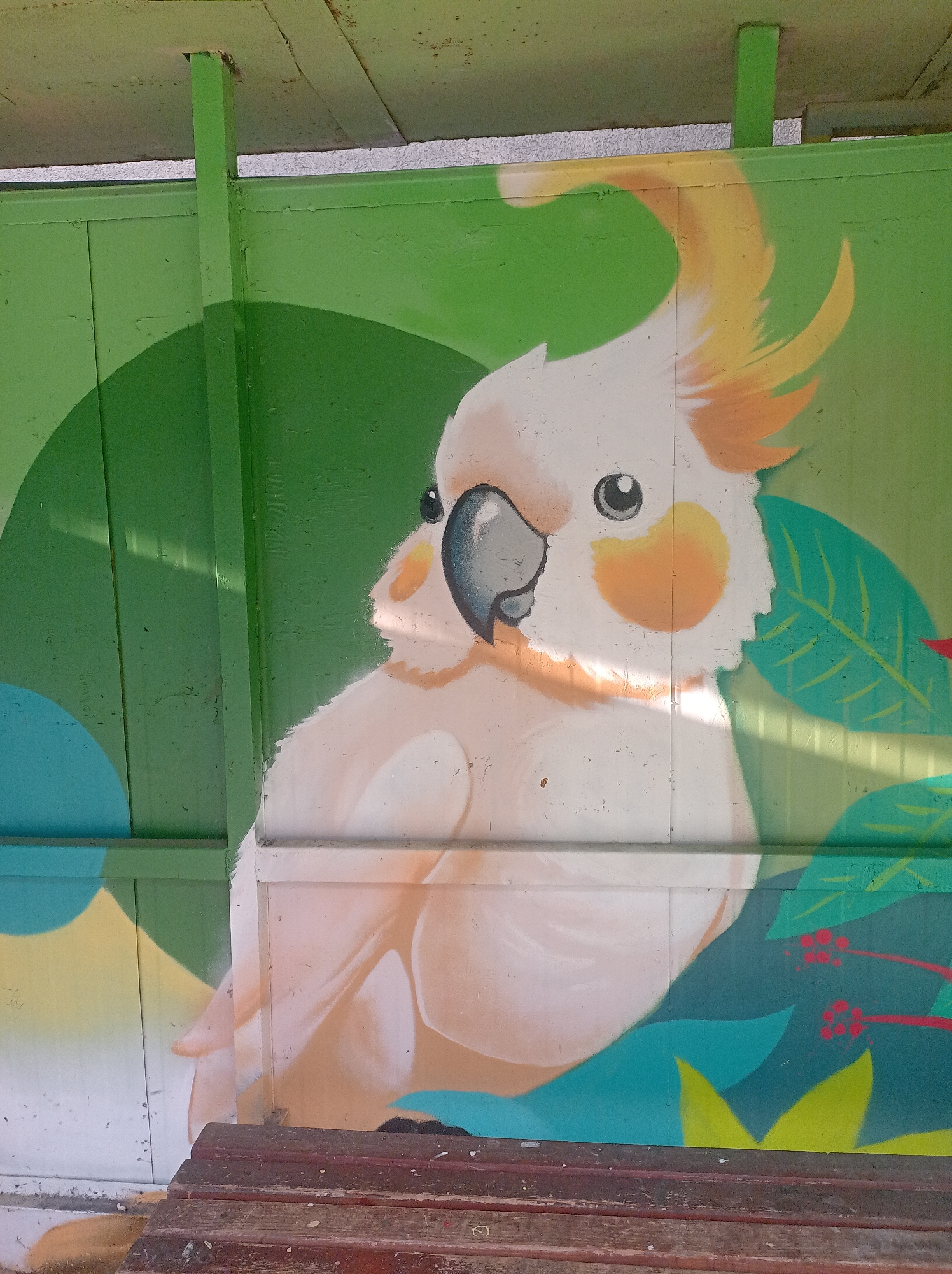 This is a medium-angle, close-up shot of a graffiti mural of a cockatoo on a green, weathered, possibly metal wall. The mural depicts the head and upper chest of a white cockatoo with orange and yellow accents on its crest and cheeks. The bird has a black beak and eye. Behind the cockatoo, there are abstract shapes and leaves painted in various shades of green and blue. A wooden bench is visible in the foreground, partially obscuring the bottom of the mural. The lighting suggests it is daytime, with shadows cast across the wall. The mural appears to be located outdoors, possibly in a public space like a park or street.