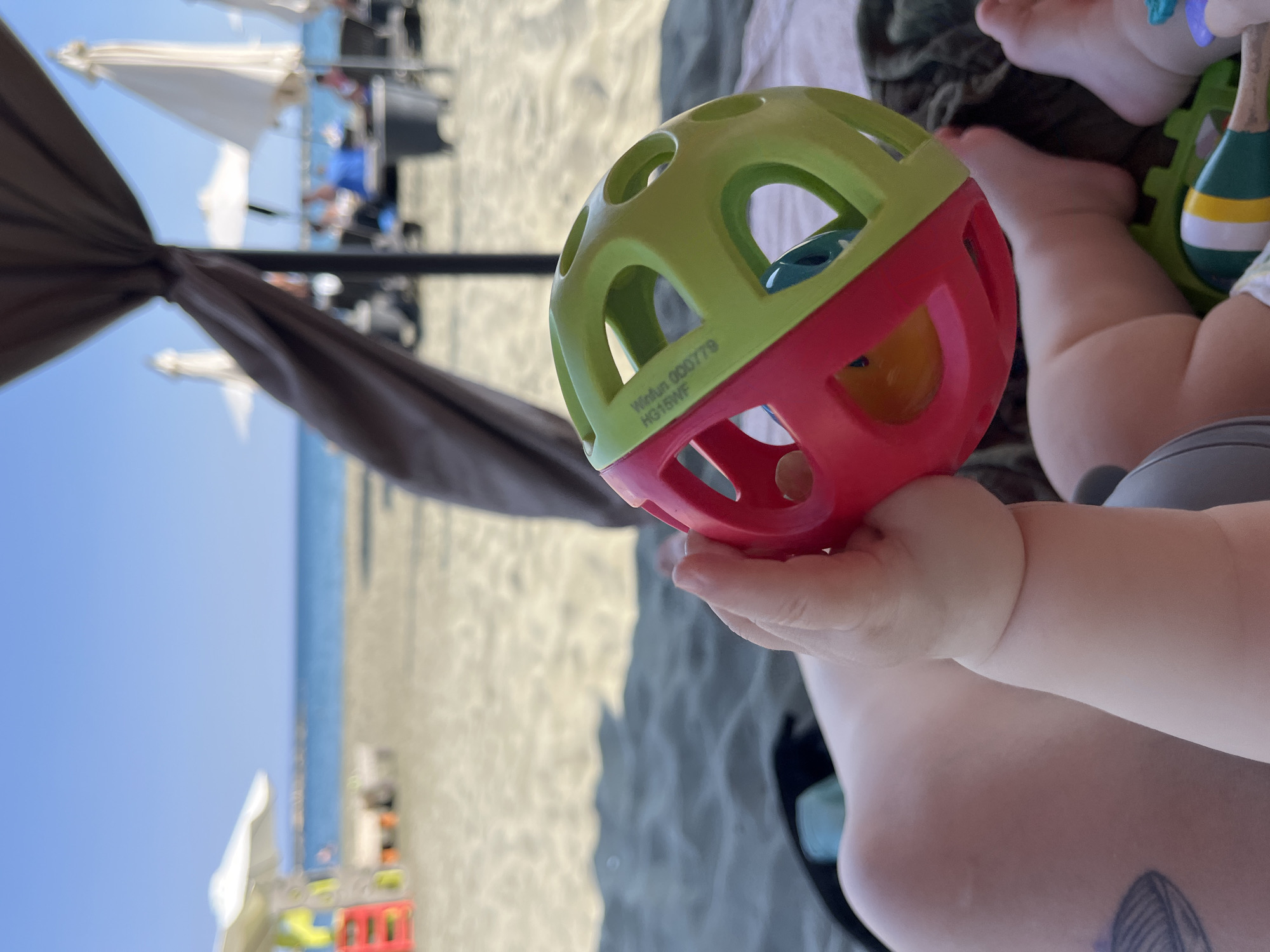 In Agios Tychona Community, Cyprus, a baby's hand holds a red and green rattle ball on a sandy beach. In the background, the calm sea meets a clear blue sky, with beach umbrellas and lounge chairs scattered across the sand. The scene evokes a sense of relaxed vacationing.