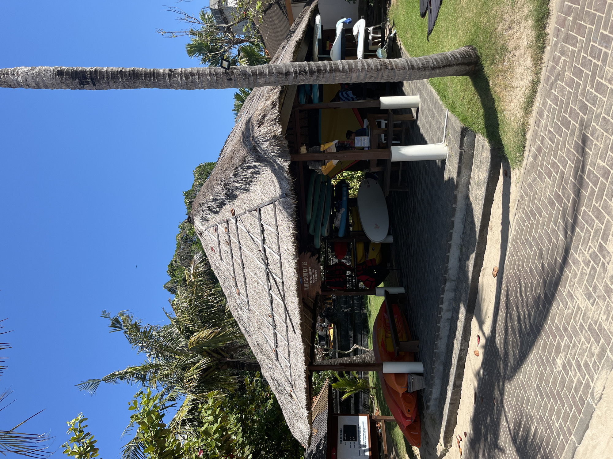 An outdoor water sport center is situated on a sandy beach in Kuta Selatan, Indonesia. A thatched-roof pavilion houses various water sports equipment, including kayaks, surfboards, and life vests, neatly organized on shelves. Orange kayaks are stacked near the entrance of the pavilion, and a wooden bench is placed inside. Adjacent to the pavilion, a sign displays the "Tide Schedule," providing information on high and low tide timings. The ground is a mixture of sand and brick paving, with palm trees and lush greenery surrounding the area. The sky is clear blue, indicating a sunny day. The scene appears to be in a tropical coastal region.