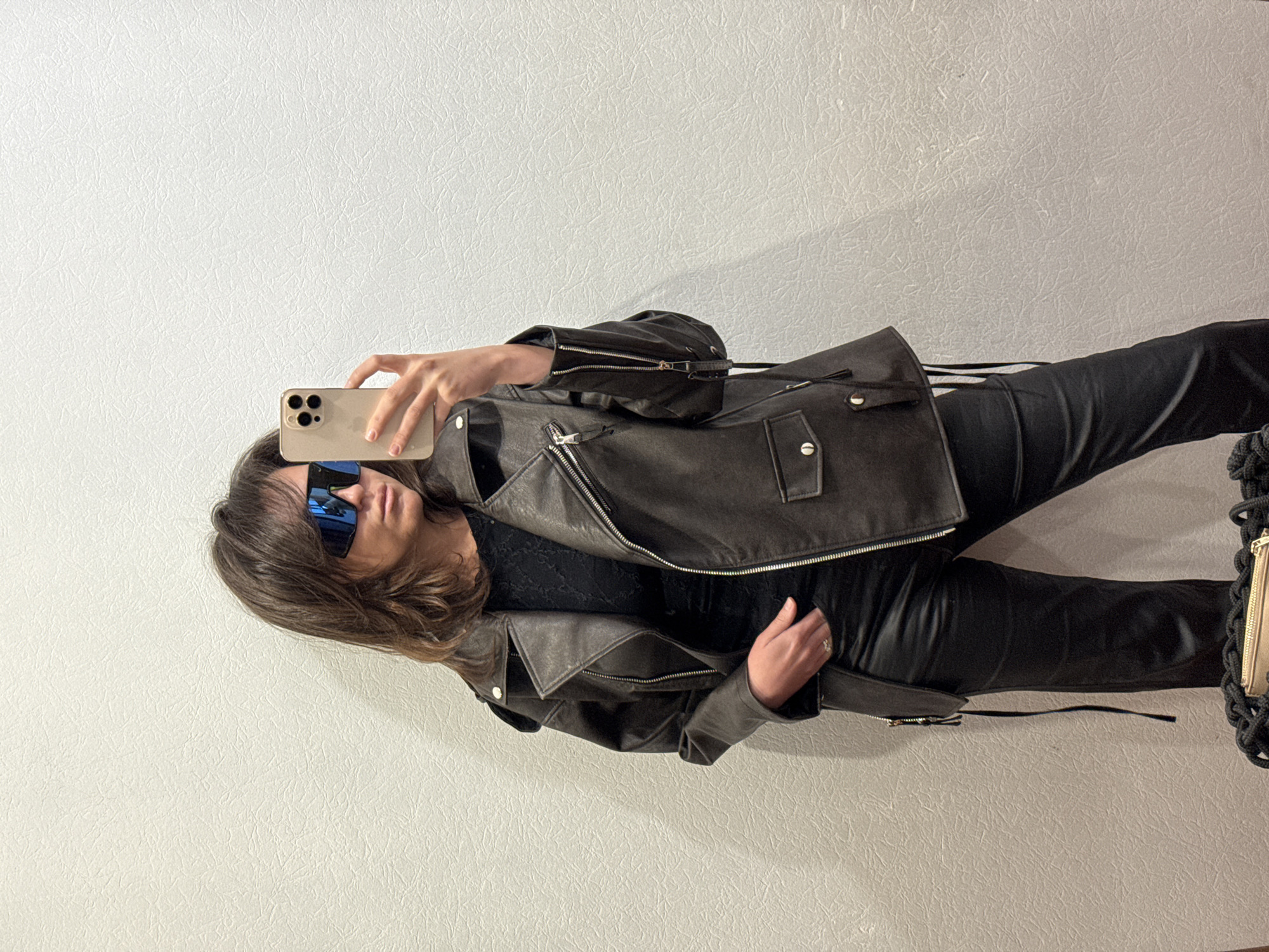 A person wearing a dark leather jacket and pants is taking a selfie with a beige smartphone. The person is wearing sunglasses with blue reflective lenses. The background appears to be a textured white wall. The presence of the location of the city of Krasnoyarsk, Russia, is not visually verifiable.