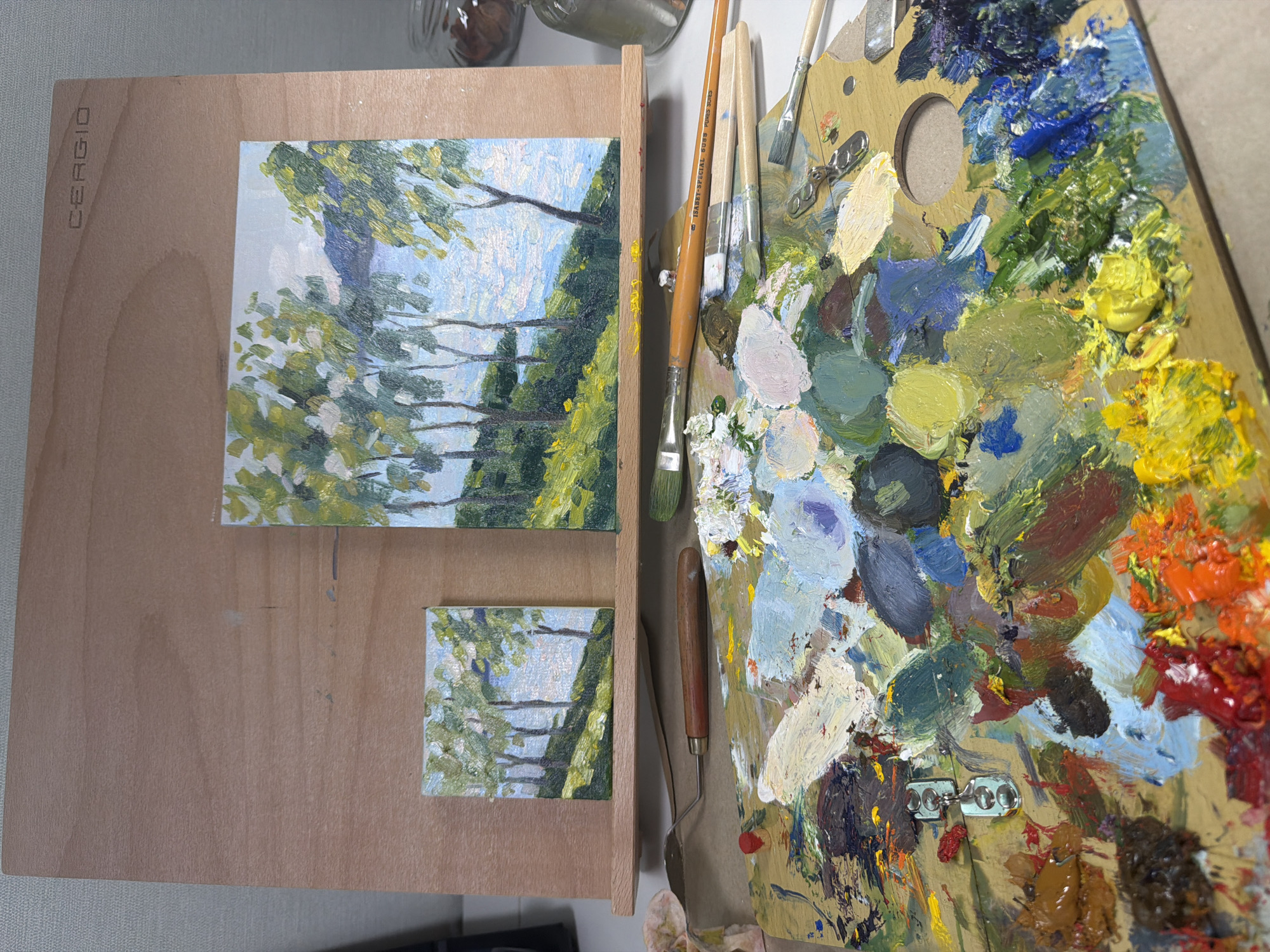 The image shows a painting setup on a wooden easel and palette. Two small, square landscape paintings, depicting trees and water, are propped up on the easel. The larger painting on the right features trees on a grassy hill overlooking a body of water. The smaller painting on the left is a miniature version of the same scene. In the foreground, a wooden artist's palette is covered in a vibrant array of oil paints, with several brushes and a palette knife scattered around it. The background is a simple, light-colored wall. There is no indication of the city of Dongducheon-si, South Korea, or any people present in the image.