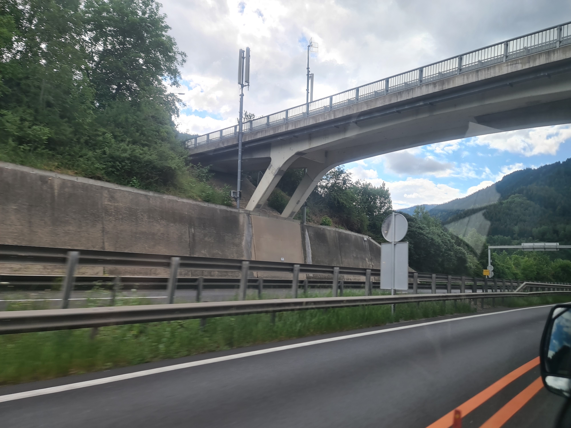 This image was taken from a vehicle traveling on a highway near Leoben, Austria, with a focus on an overpass. The overpass, constructed of concrete and steel, spans the highway. On the left side of the highway, a steep, vegetated embankment rises, topped by a concrete retaining wall. Two tall poles with communication equipment are visible on the embankment near the overpass. On the right side of the highway, the landscape transitions to forested hills. The road itself is asphalt with white and orange lane markings. A metal guardrail separates the highway lanes from the embankment. The sky is partly cloudy, with a mix of grey clouds and blue sky. The image captures a typical highway scene, emphasizing the infrastructure and natural surroundings of the area.