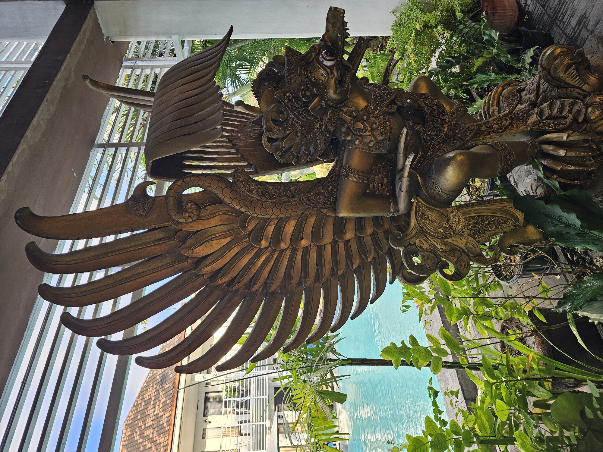 A bronze statue of a Garuda, a mythical bird-like creature, stands prominently in the foreground. The statue is highly detailed, depicting the Garuda with large, feathered wings, a human-like torso, and a bird's head with a beak. It is adorned with intricate ornaments and a headdress.
Behind the statue, a bright blue swimming pool is visible, lined with a light-colored tiled edge. To the left of the pool, a building with a tiled roof and white balconies is partially visible, suggesting it is a hotel or resort. Lush green plants and palm trees surround the pool area and the statue, creating a tropical ambiance.
The scene is framed by a white structure with horizontal louvers overhead, which allows sunlight to filter through, casting shadows. The environment suggests a warm, outdoor setting, likely in a tourist-oriented location. While the image does not explicitly state the location, the architectural style, lush vegetation, and the presence of a swimming pool are consistent with many resorts and hotels in Denpasar, Indonesia, a popular tourist destination known for its cultural heritage and tropical beauty. The statue itself is characteristic of Balinese art and mythology. There are no people or animals in the image.