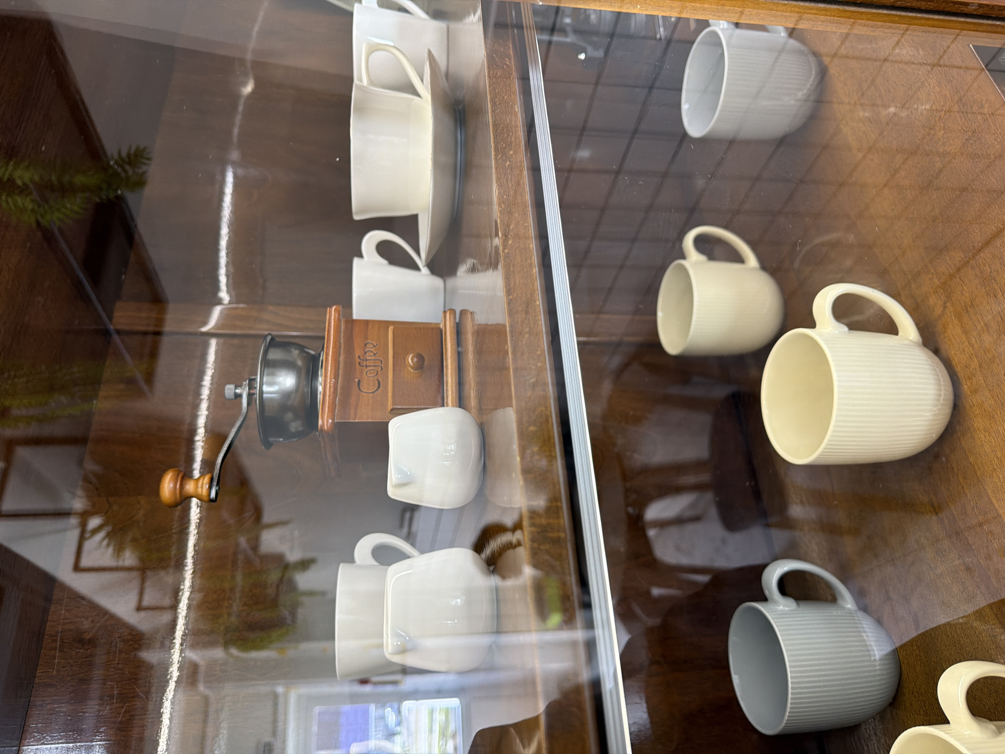 The image shows a display case with two sections. The left section contains a stack of white cups and saucers, a manual coffee grinder, and a small, white, cat-shaped container. The right section displays several white mugs, some with horizontal ribbing. The mugs vary in tone from off-white to gray. The environment is likely a shop or museum. The location is Chaikovsky, Russia, but there is no information to verify that in the image.