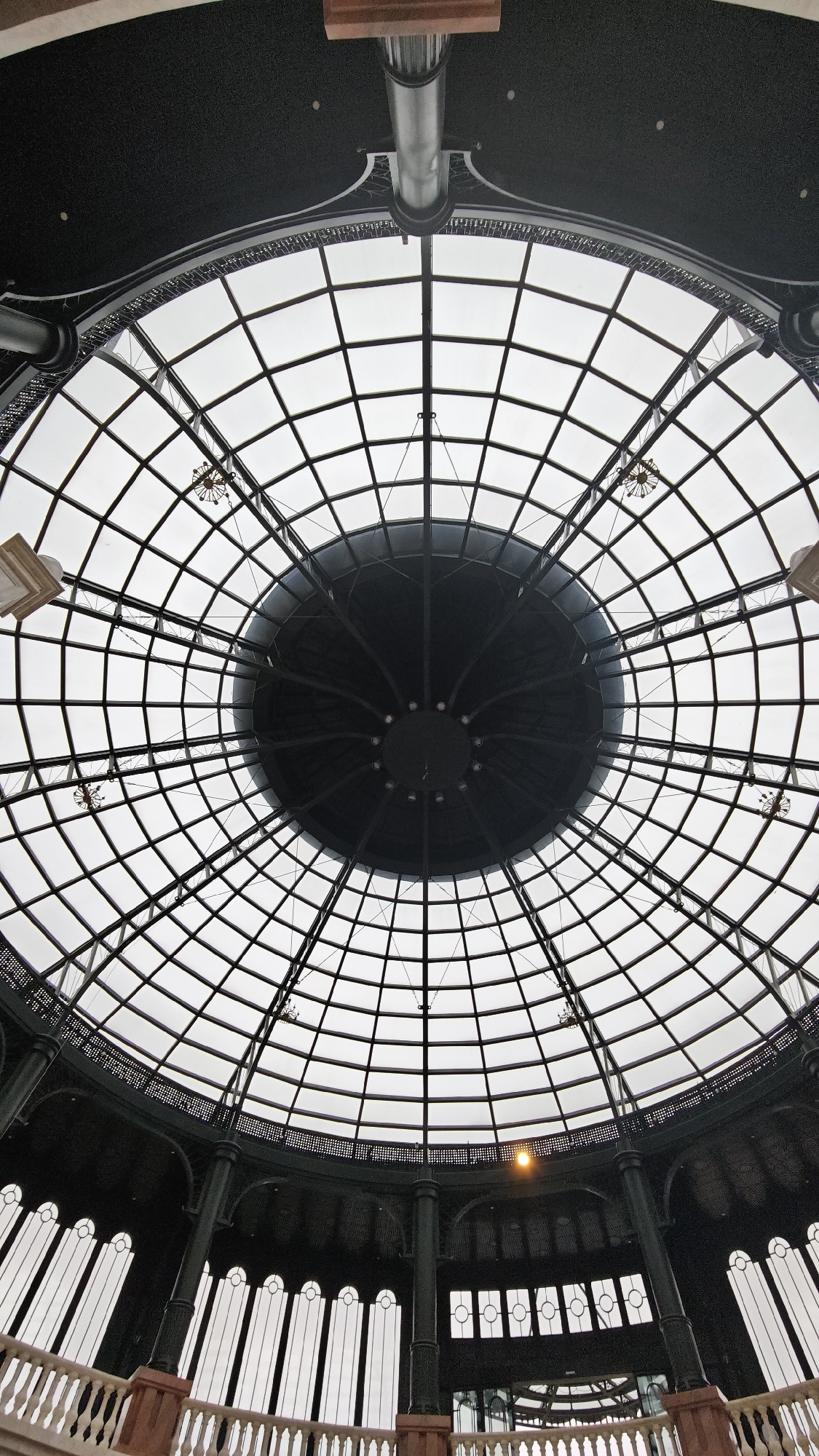The image presents an upward perspective into a grand architectural interior, showcasing an elaborate glass dome and the structure beneath it, located in Da Nang, Vietnam. The dominant feature is a large, circular glass dome composed of numerous rectangular and trapezoidal panels framed by a dark, intricate metal grid. At the dome's zenith, a prominent, dark, circular structure with radiating spokes and small embedded lights forms a central element, possibly a large light fixture or decorative cap. Several smaller, ornate chandeliers or light fixtures are suspended at various points from the metal framework of the dome.
Below the expansive skylight, a dark, decorative frieze or band wraps around the circumference, detailed with patterned metalwork. Further down, sturdy, dark-colored columns with classical elements rise to support the structure. On the right side, a single warm-toned light fixture glows against the dark background of the wall. The bottom of the image reveals a partial view of a balustrade or railing, featuring reddish-brown newel posts and lighter-colored balusters, indicating a balcony or mezzanine level. Behind this railing, a series of tall, arched window-like openings, appearing uniformly white, are set into the dark lower wall.
The lighting through the dome is bright and diffused, suggesting an overcast sky or well-distributed artificial lighting. There are no people visible, and no discernible text is present in the photograph. The scene conveys a strong sense of architectural detail and grand interior design.