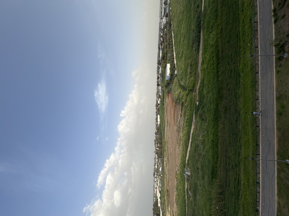 An elevated long shot depicts a semi-urban landscape in Yavne, Israel, under a partly cloudy blue sky. The upper half of the image features a clear blue sky transitioning to scattered white cumulus clouds along the horizon line, particularly prominent on the left. The midground consists of an expanse of mixed terrain: a large, irregular brown field occupies the central-left, bordered by vibrant green fields and vegetation to its right and foreground. Dirt paths traverse these green areas, and a small, white, possibly utilitarian structure is visible within the green field in the center-left. Further in the background, a density of residential and industrial buildings, interspersed with trees, defines a cityscape along the horizon. A white-roofed building is present in the mid-right within a green section. In the foreground, a dark asphalt road or pathway stretches horizontally across the bottom of the frame, separated from the green fields by a dark, low-height guardrail. Cracks are visible on the road surface. A single individual, dressed in light-colored clothing, walks from right to left on this paved path. A dark grey streetlamp pole stands upright on the right side of the path, extending upwards from the lower frame edge.