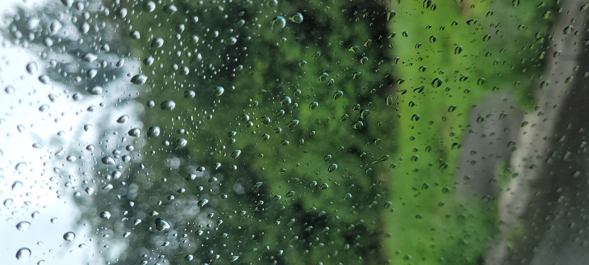 This image is a close-up view of raindrops on a window pane. The background is blurred, showing greenery and what appears to be a path or road. The overall impression is one of a rainy day, likely viewed from indoors. There is no indication of the location being in Dietzenbach, Germany.