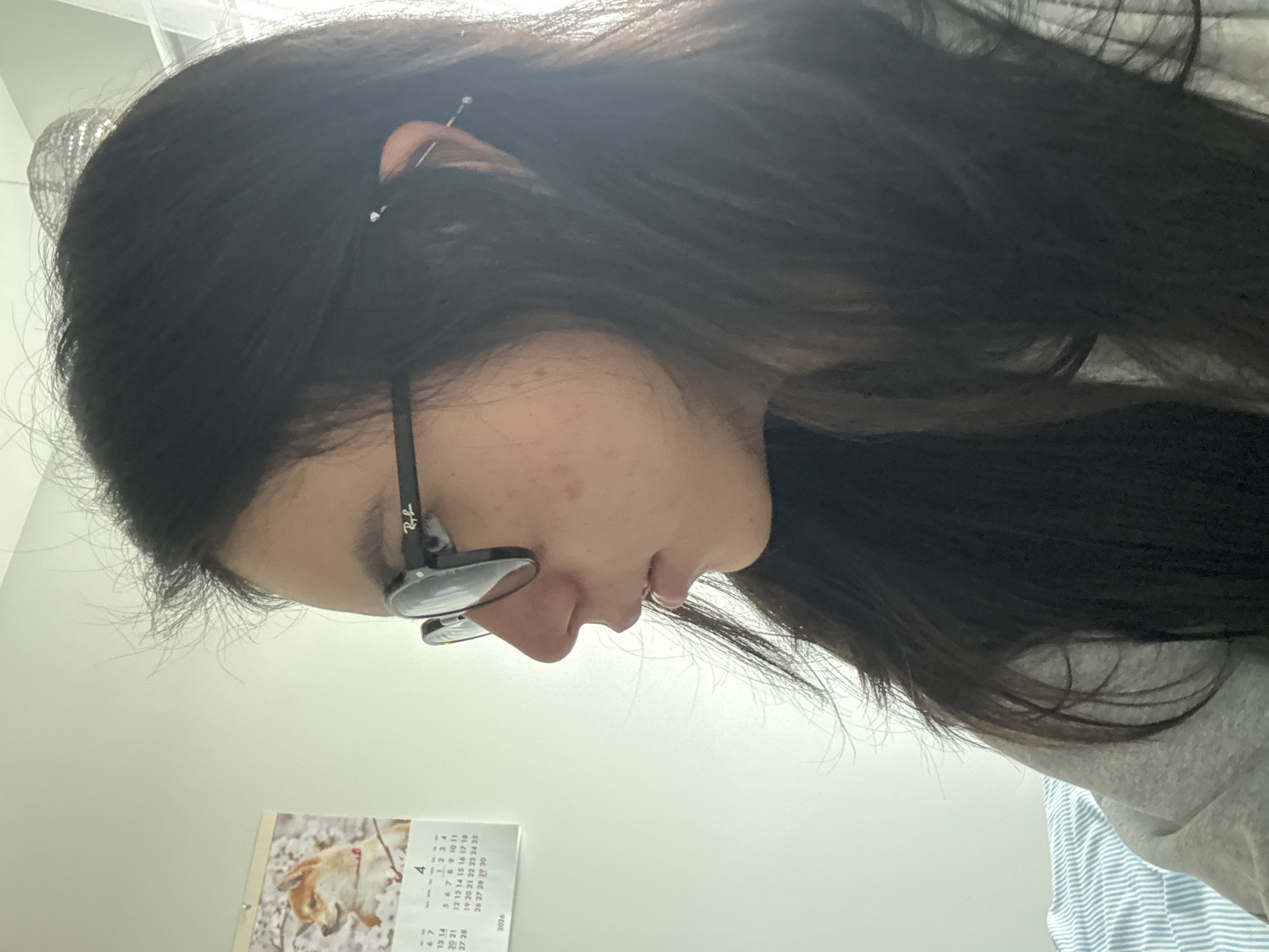A woman with long, dark hair and glasses is looking down. She is wearing a light gray top. A calendar is visible on the wall behind her, featuring an image of a dog. The calendar is set to April 2026. The scene is indoors. There is no indication of the location being Austin, United States of America.