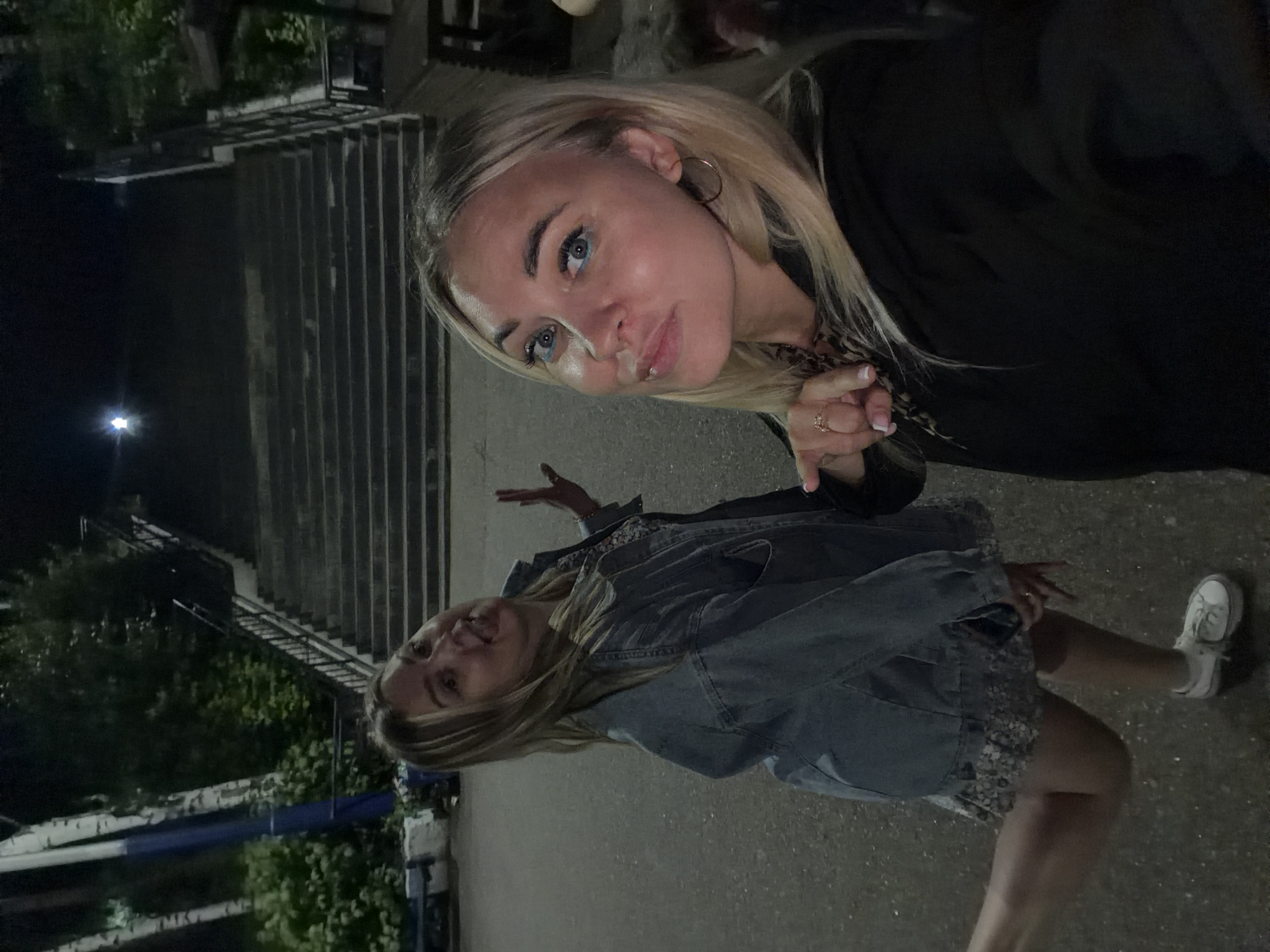 Two young women pose for a selfie at night in front of a concrete staircase in Chaikovsky, Russia. The woman in the foreground, wearing a black hoodie, smiles towards the camera with blue eyeliner. She is wearing a ring on her left hand. The woman behind her, wearing a denim jacket and a floral skirt, sticks her tongue out at the camera and holds her left hand up. The background shows a dark, tree-lined area with a long, wide staircase leading upwards. The lighting suggests an outdoor public space.