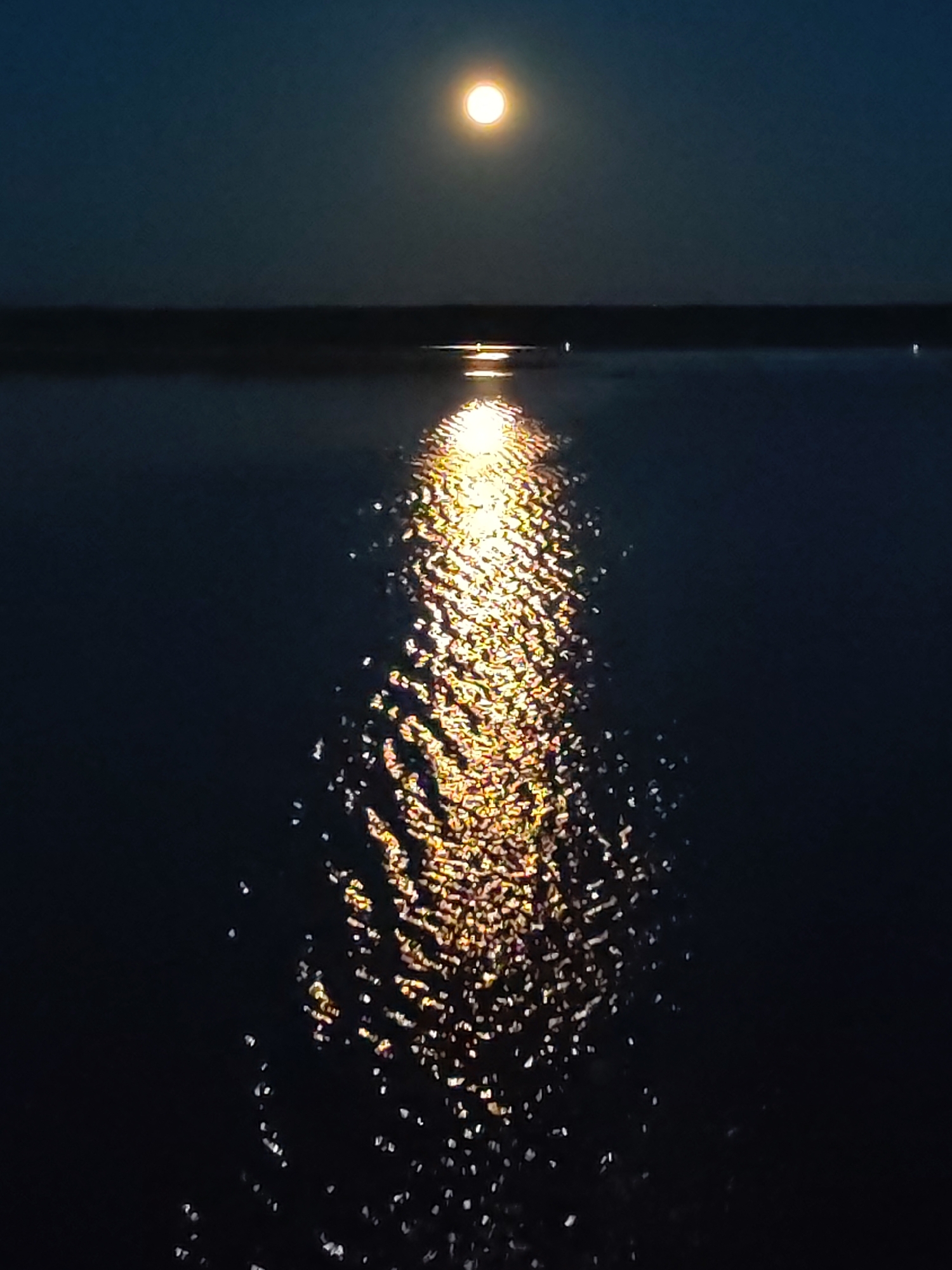 The image depicts a body of water reflecting moonlight. A bright, round moon is visible in the sky, casting a shimmering, golden path across the water's surface. The surrounding environment appears dark, with a treeline visible in the distance. The location is provided as Irdomatka, Russia. No people, subjects, objects, actions, or interactions are apparent in the scene, other than the natural light reflecting on the water.