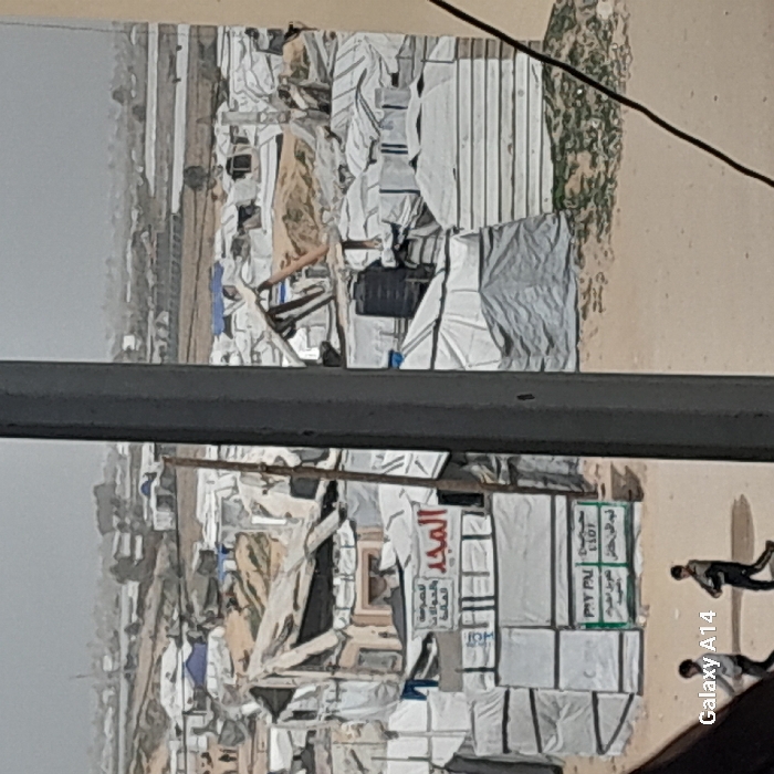 The image displays a densely packed, temporary settlement within the city of Khan Yunis, Palestinian Territory, viewed from an elevated perspective. Numerous makeshift shelters, predominantly constructed from white and light-colored tarpaulins, plastic sheets, and fabric, are arranged closely together on sandy or dirt ground. Several structures exhibit signs of damage or partial collapse, with exposed frameworks and torn materials.
In the midground, a multi-sectioned shelter features a white banner with prominent red Arabic text, "المجد" (Al-Majd), accompanied by smaller black Arabic script. Below this, a green sign displays "PAY PAL" alongside additional Arabic text. A dark, cylindrical water tank is positioned on the roof of an adjacent shelter. A wooden pole stands vertically near the signs.
The terrain consists of barren, light-brown earth with sparse patches of green vegetation. In the lower-left foreground, two individuals, dressed in dark clothing, walk from left to right on a dirt path. One appears to be carrying an object in his right hand. A thick, dark grey vertical beam partially obscures the right side of the foreground, with a black cable running diagonally across the lower right. The background shows a continuation of the encampment, fading into a hazy horizon under an overcast sky.