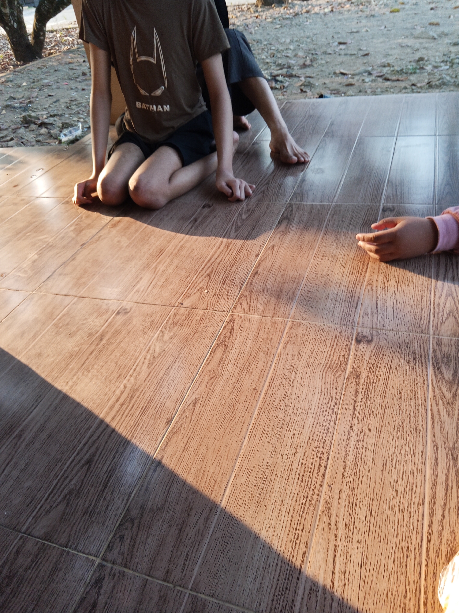 The image shows a close-up view of two children sitting on a tiled floor outdoors. The child on the left is wearing a brown t-shirt with a Batman logo and black shorts. Their knees are bent, and they are resting their hands on the floor. The child on the right is partially visible, with their hands resting on the floor. The floor is made of brown, wood-patterned tiles. Sunlight casts a shadow across the tiles. The background is out of focus, showing some trees and dirt. The location is the outdoor area of a home in Lubuklinggau, Indonesia.