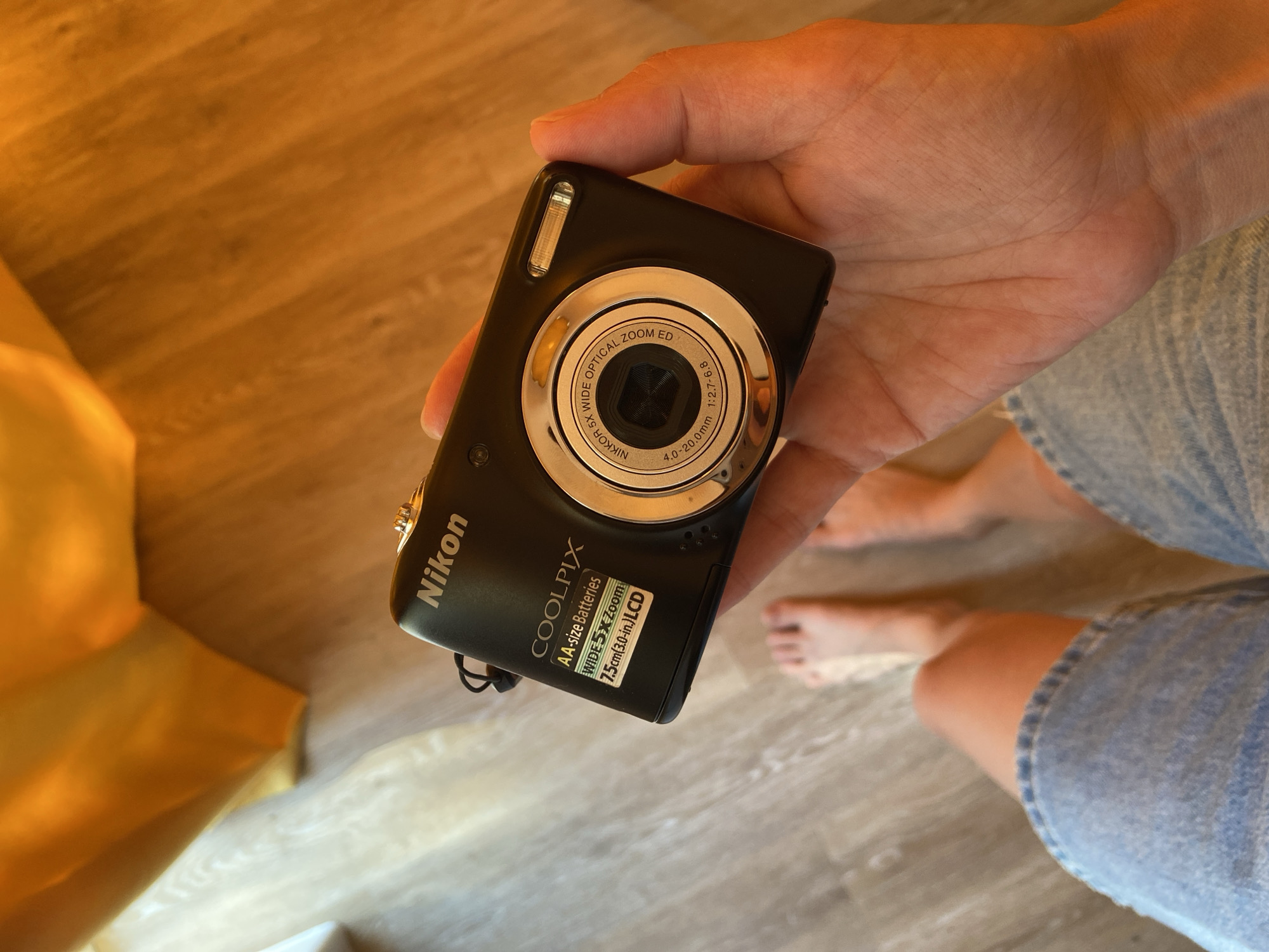 A person is holding a black Nikon Coolpix digital camera in their hand. The camera has a silver ring around the lens with "NIKKOR 5X WIDE OPTICAL ZOOM ED" and "4.0-20.0mm 1:2.7-6.8" printed on it. The camera's lower portion has a label that reads "COOLPIX," "AA-size Batteries," "WIDE 5X Zoom," and "7.5cm(3.0-in.) LCD." A section of a yellow-colored cloth is seen to the left of the camera. The person's legs and feet are visible in the background, with the person wearing light blue denim shorts or pants. The floor appears to be light-colored wood. The city of Tyumen, Russia is not discernible from the information in the image.