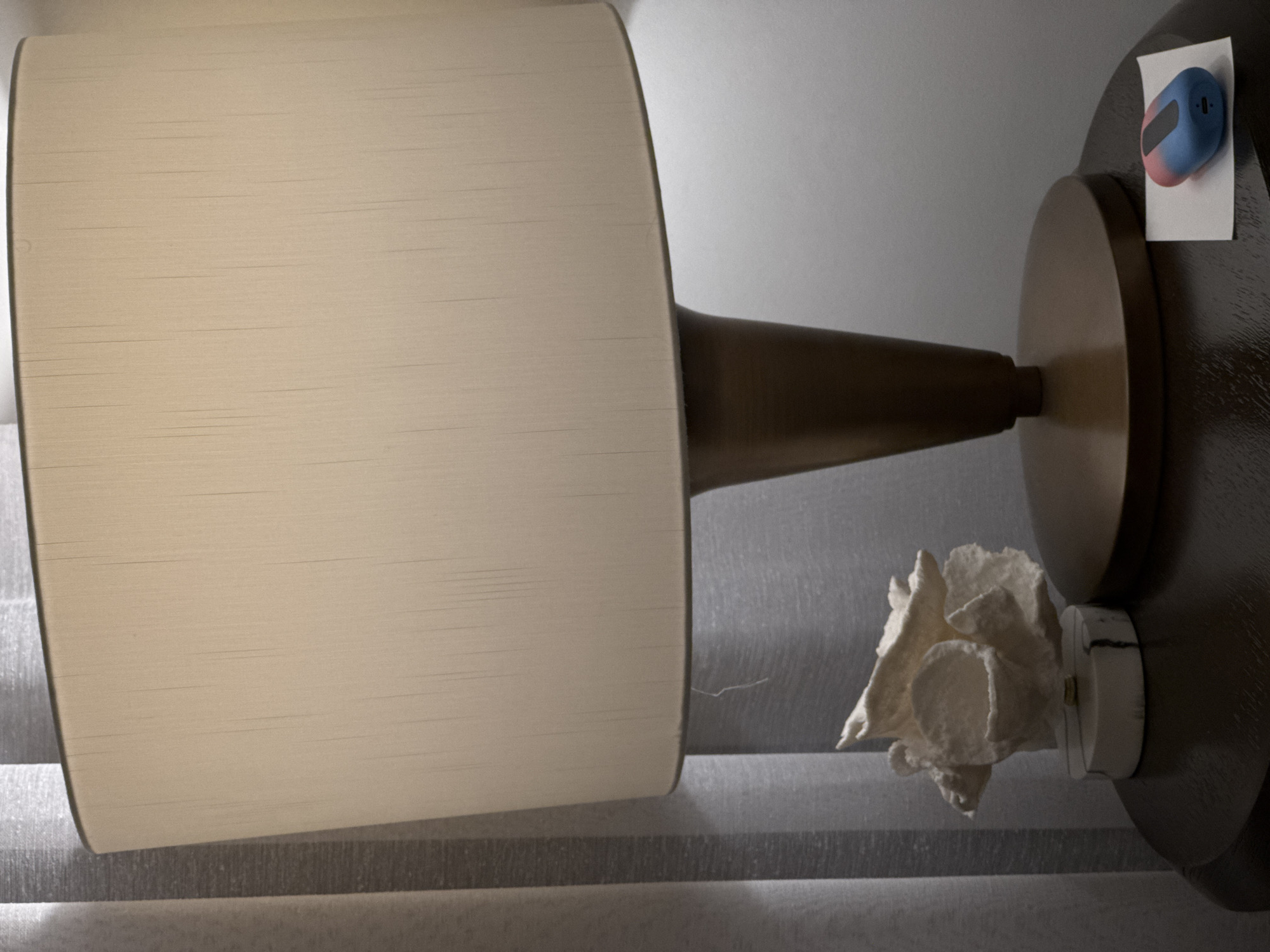A close-up shot of a lamp on a table, presented from an unusual perspective. The lampshade, the most prominent element, is large and off-white in color. Its position indicates that the camera is either positioned low or the lamp is angled downwards. Beside the lamp, slightly obscured, there's a decorative piece that could be a white flower or sculpted paper. Attached to the lamp's base is a blue and pink electronic device secured with a piece of white paper. The table appears to be a dark, polished wood. The background is a curtain. The location and city are unknown.