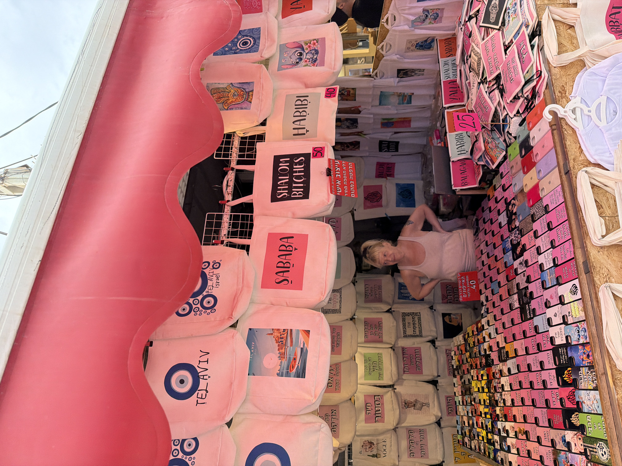 The image shows a narrow shop in Tel-Aviv, Israel, filled with merchandise, including tote bags and phone cases, likely aimed at tourists. A fair-skinned woman with blonde hair is visible inside the shop. The shop's entrance is framed by a pink arch. The bags feature local phrases in Hebrew and English, and city-related designs, like an evil eye. Phone cases are arranged by color, with various designs. Price tags are displayed. The overall environment seems crowded.