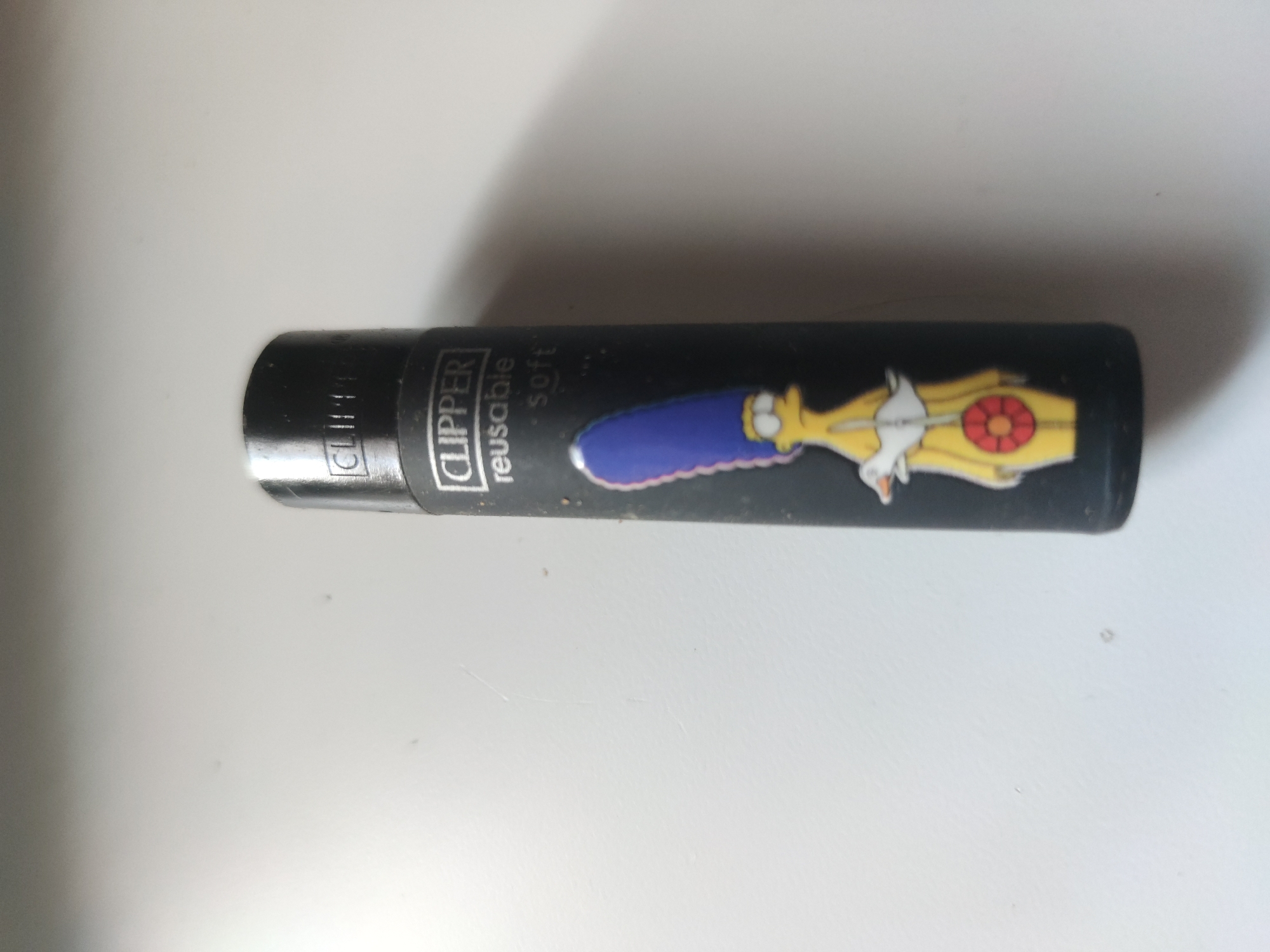 A black Clipper lighter with a depiction of Marge Simpson on it lies on a white surface. The lighter is positioned horizontally in the image, with the top end slightly to the left. "CLIPPER" and "reusable soft" are printed on the lighter. The Simpson character is shown riding a yellow submarine.