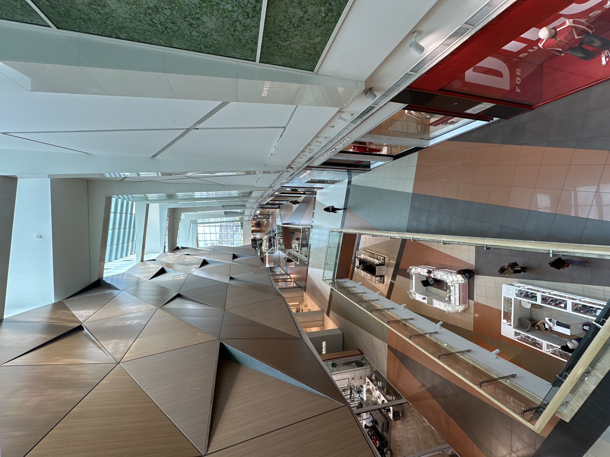 The image presents an overhead view of a modern shopping mall interior. The composition is dominated by an angled perspective, showcasing the mall's multi-level layout. The left side of the frame features a distinctive wall design composed of geometrically arranged metallic triangles. Windows are apparent on the left, indicating an exterior view.
The right side of the image reveals the retail spaces within the mall, including storefronts with glass facades. There are people visible on the floor, possibly shoppers, and displays with items for sale are visible.
Details include:
* Flooring patterns with a mix of grey, beige, and brown tones.
* Overhead lighting fixtures.
* Red storefront with the word “DIESEL.”
* Reflections on glass surfaces.
Although the image does not state the location, based on the style and architecture, it is likely within a contemporary shopping center such as the Paradise Center in Sofia, Bulgaria. However, this location cannot be confirmed without additional information.