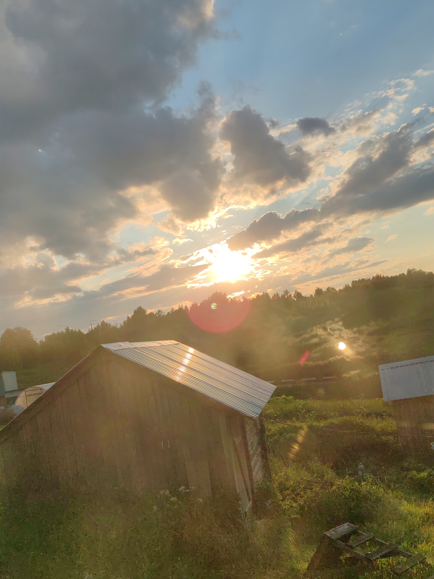 The image depicts a rustic outdoor scene, likely in a rural area of Чухлэм, Russia, during a sunset. The foreground is dominated by a weathered wooden structure with a corrugated metal roof that reflects the golden light of the setting sun. Tall grass and some wildflowers grow around the base of the structure.
In the middle ground, a dense line of trees forms a dark silhouette against the sky. The sky itself is a dramatic canvas of clouds, with the sun breaking through them, casting a warm, hazy glow over the landscape. Rays of sunlight are visible, and there are lens flares in the shape of red circles, indicating the sun's direct presence. In the distance, a slightly elevated area with sparse vegetation is visible, also bathed in the sunset's light, with a faint shimmering effect suggesting mist or atmospheric haze. Another structure with a light-colored roof is partially visible on the right side of the image. The overall impression is one of a tranquil, natural setting at dusk.