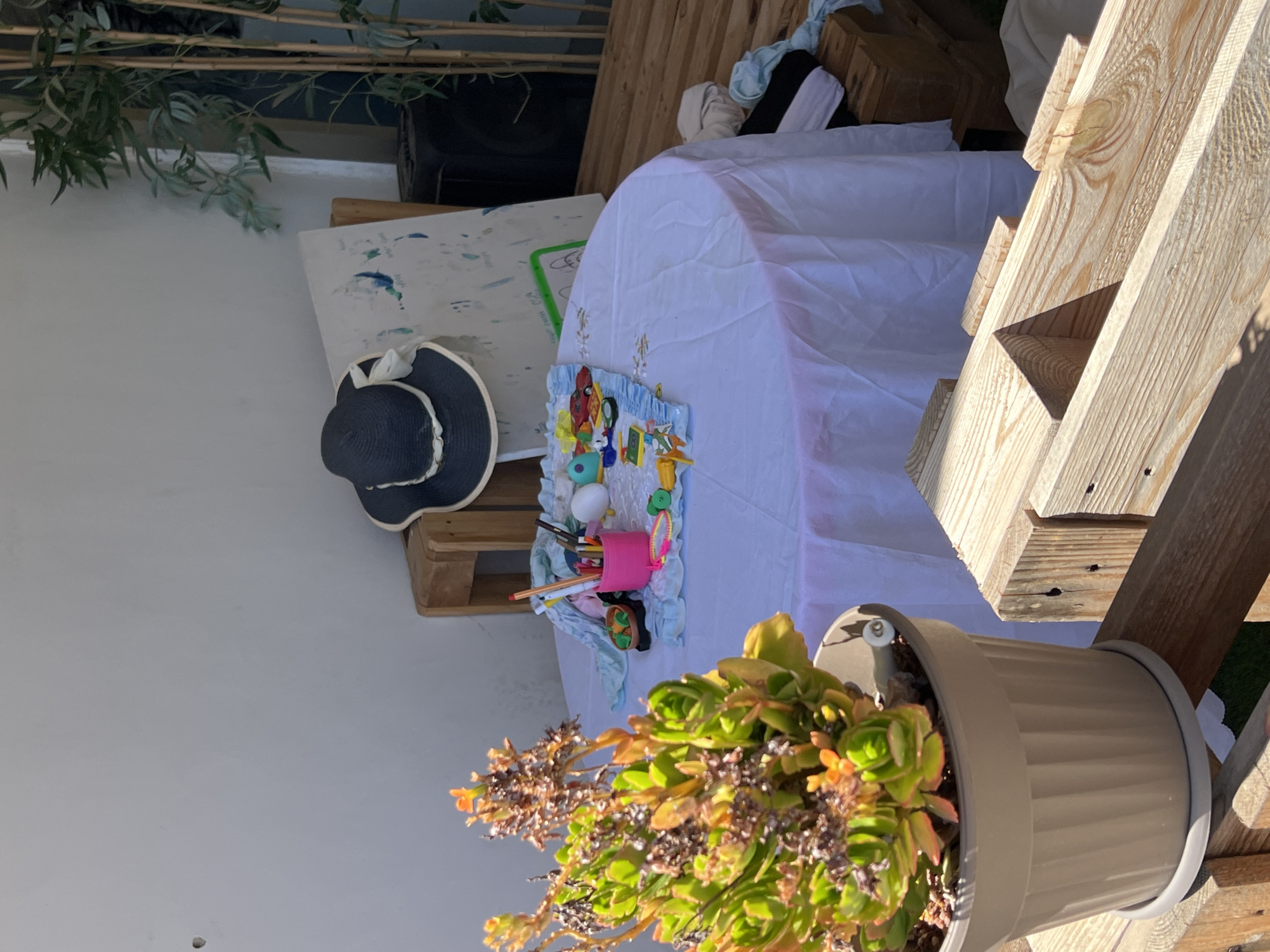 The image shows an outdoor setting in Kiryat Bialik, Israel. There is a wooden pallet table covered with a white tablecloth. On the table are art supplies including paintbrushes, paint, and a child’s craft project. A navy blue sun hat is placed on the side of the table. In the foreground, there is a potted succulent plant. The background includes a white wall and green foliage. Sunlight is present, casting shadows on the wooden structures. There are no people visible in the image.