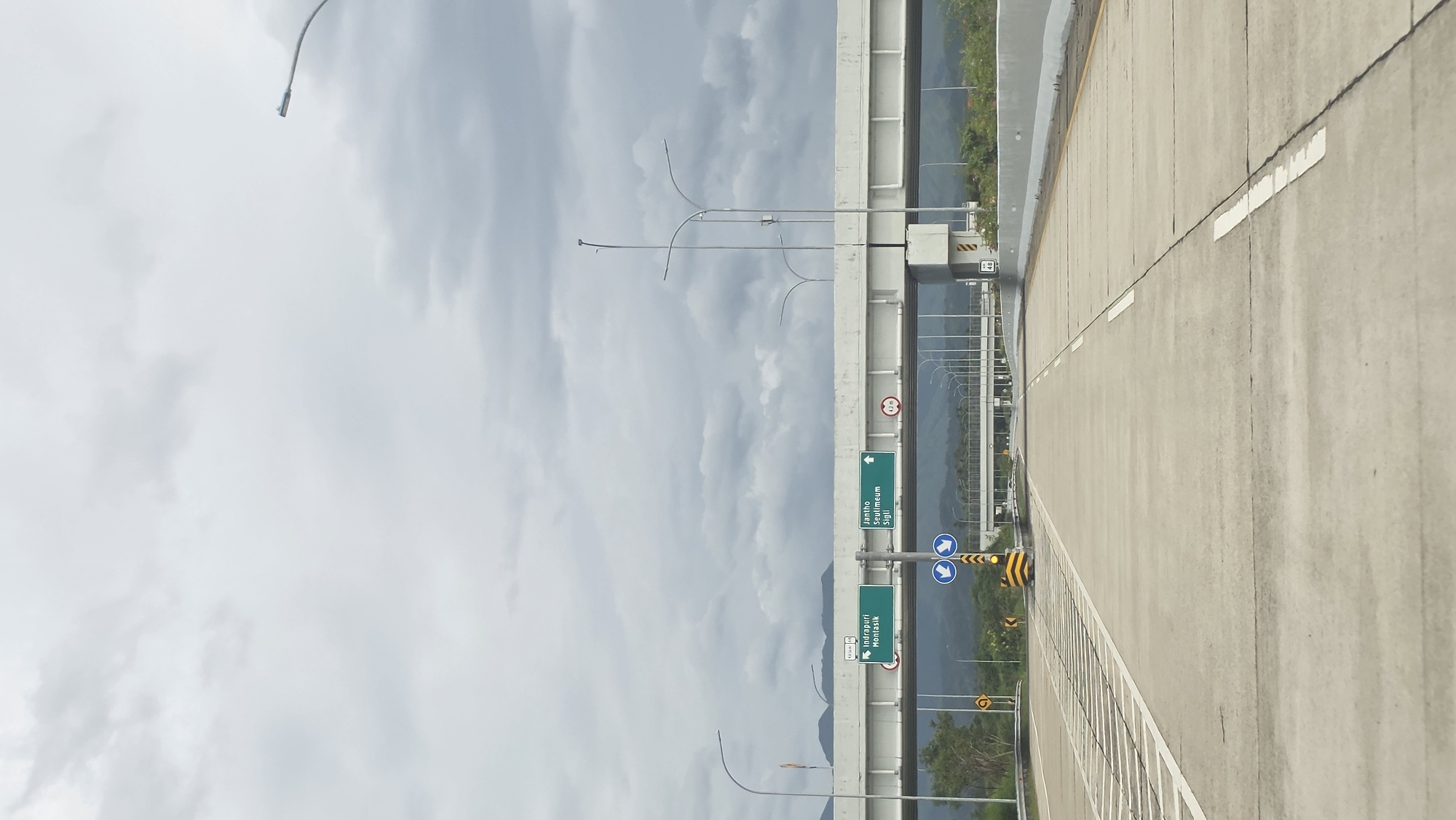 The image shows an elevated highway in Lampanahtunong, Indonesia. The road surface is concrete, and white lane markings are visible. To the left of the road, there is a concrete barrier with vertical metal supports. Signage indicates directions to Jantho, Seulimeum, Sigli, Indra Puri, and Montasik. The sky is overcast with grey and white clouds. Streetlights are present along the highway. The background consists of forested hills.